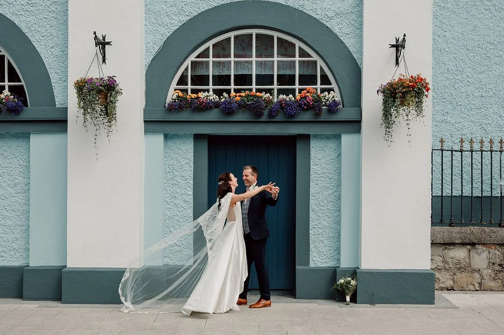 Wedding couple dancing in front of a blue building with flower boxes and hanging planters.