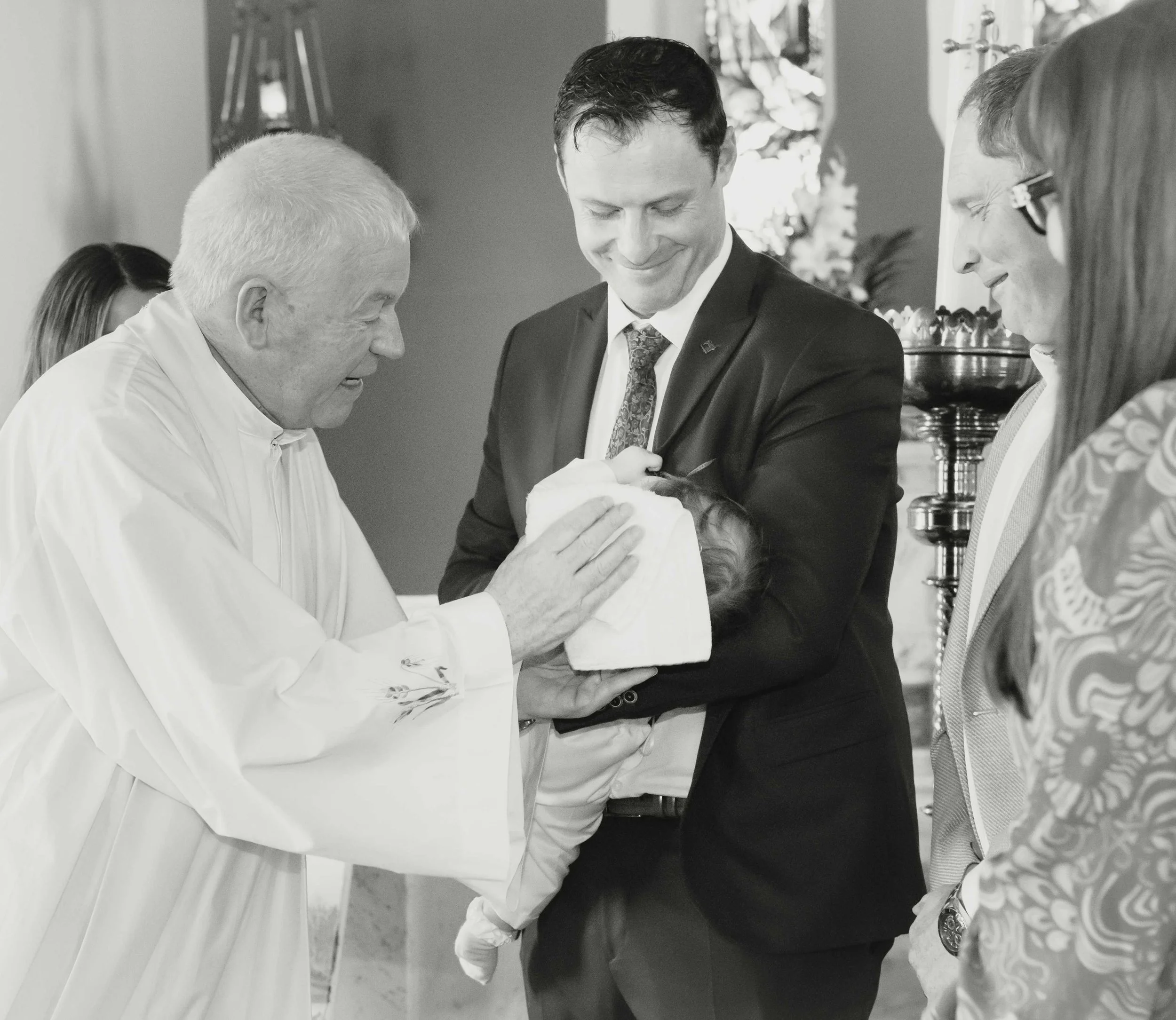 A priest baptizing a baby with a cloth held by a man in a suit, surrounded by attentive family members during a christening ceremony in Ballyheigue, Co.Kerry