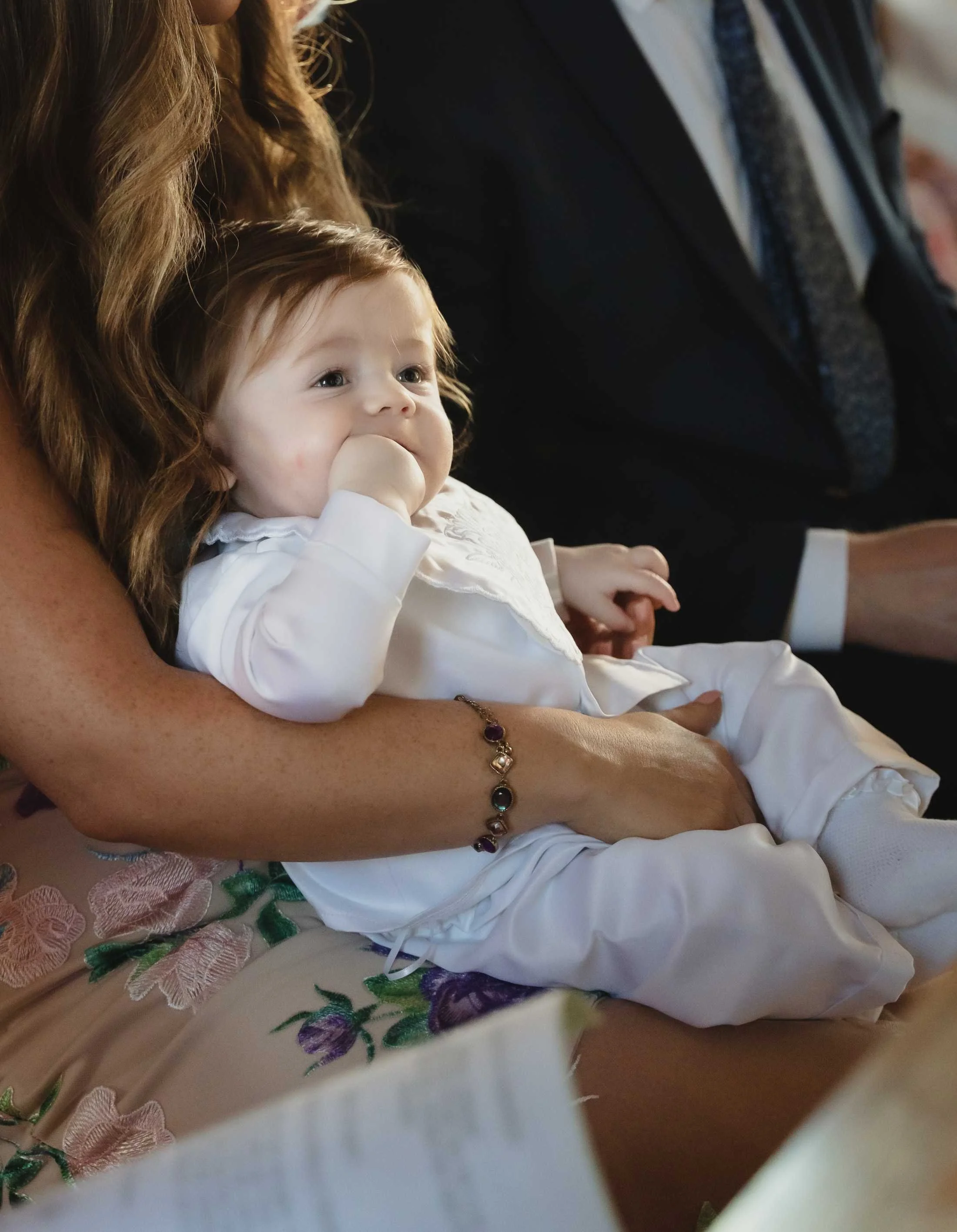 A portrait of a young child with light brown hair in a white outfit, sitting on mothers lap during his christening ceremony in Ballyheigue, Co.Kerry.