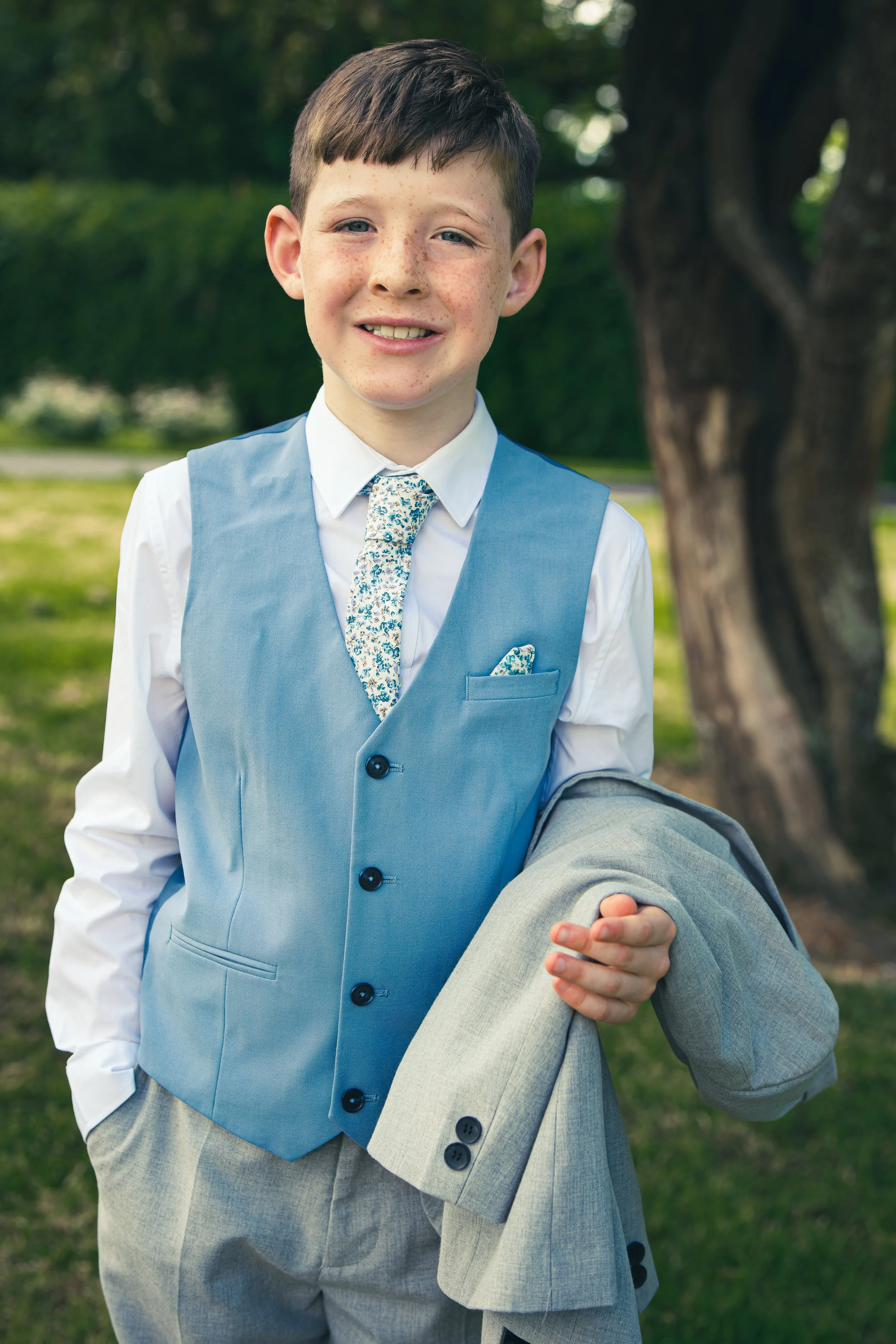 A young boy after his communion in Tralee standing in a light blue vest, white shirt, and patterned tie, holding a gray suit jacket in an outdoor park setting.