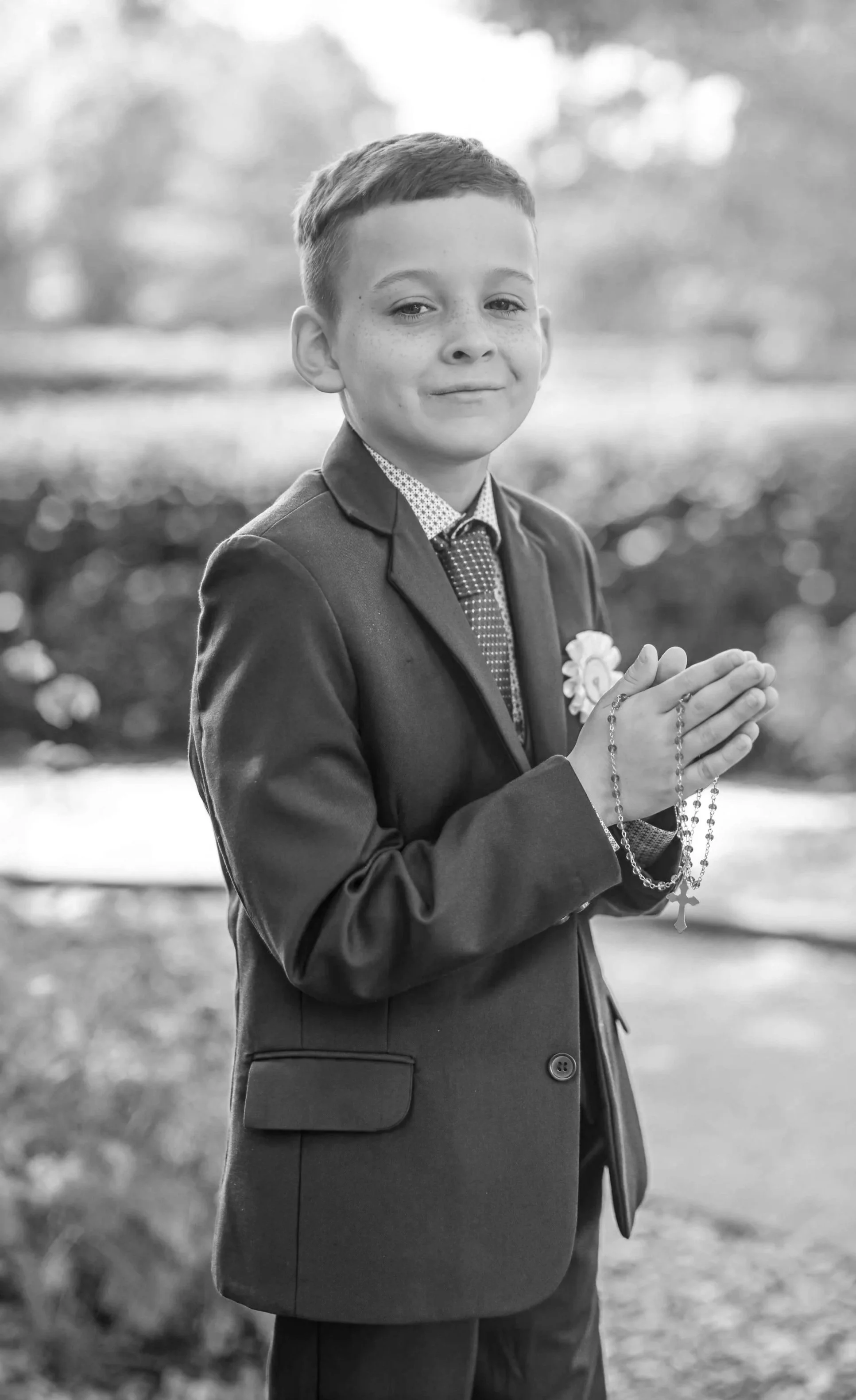 Black and white photo of a young boy in a suit holding a rosary with a cross, standing outdoors with a blurry background after his communion in Tralee