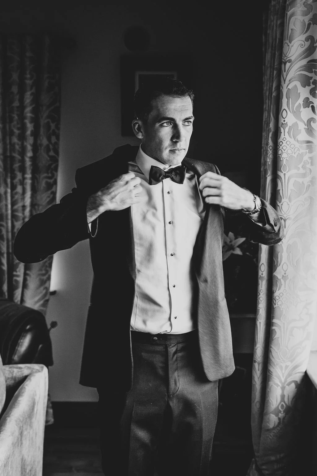 A man in formal attire, adjusting his jacket, standing in a room with patterned curtains and a window. The image is in black and white.