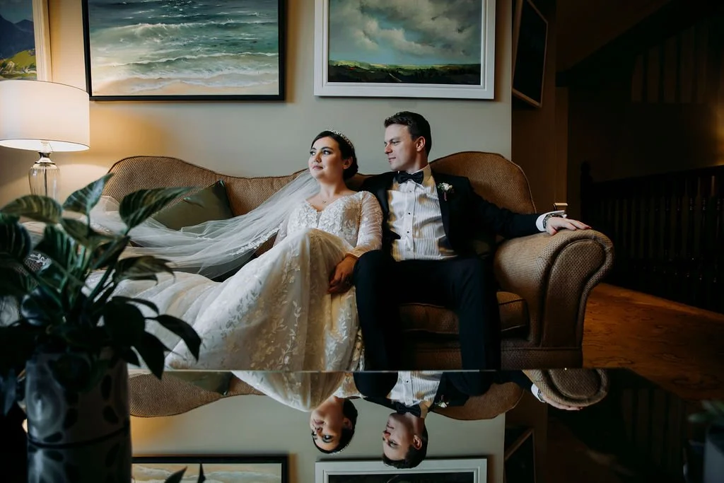 A bride and groom sitting on a beige sofa in a cozy living room, with a mirror below reflecting their image, surrounded by framed landscape paintings and indoor plants.