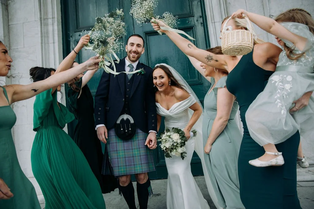 A bride and groom outside a building surrounded by bridesmaids and guests, celebrating with flowers and joyful expressions.
