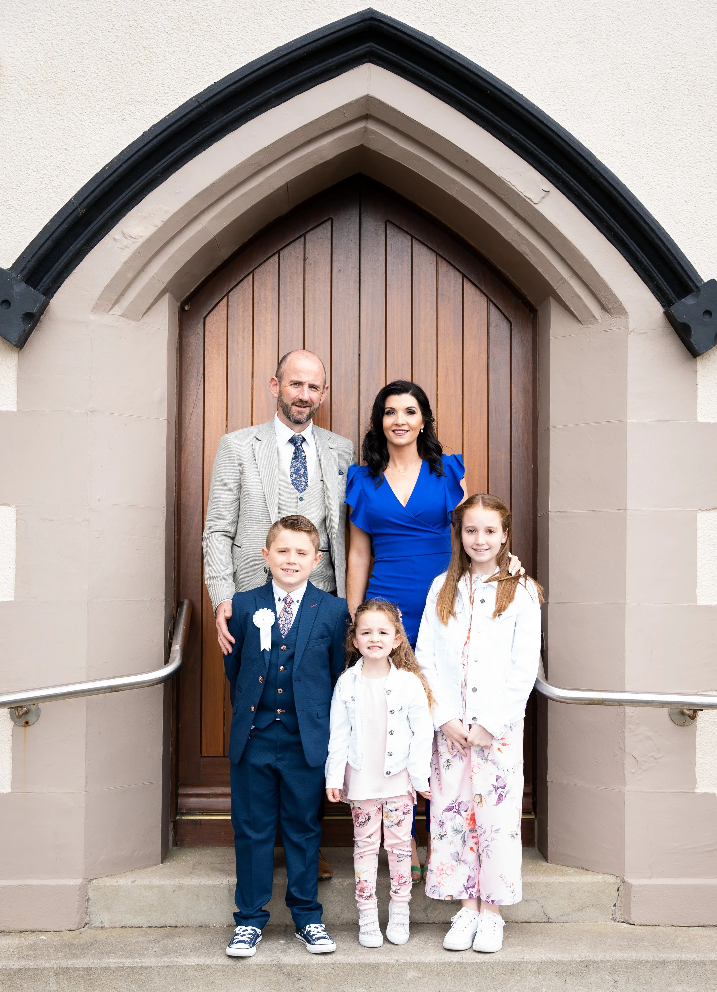 A photo of a family posing in front of Ballyheigue Church, Co. Kerry after communion ceremony.  The parents stand behind three children, all smiling. 