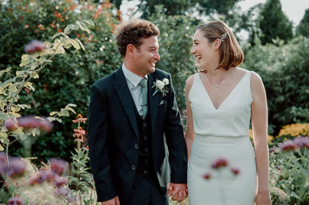 A newlywed couple holding hands and smiling at each other in a garden with colorful flowers.