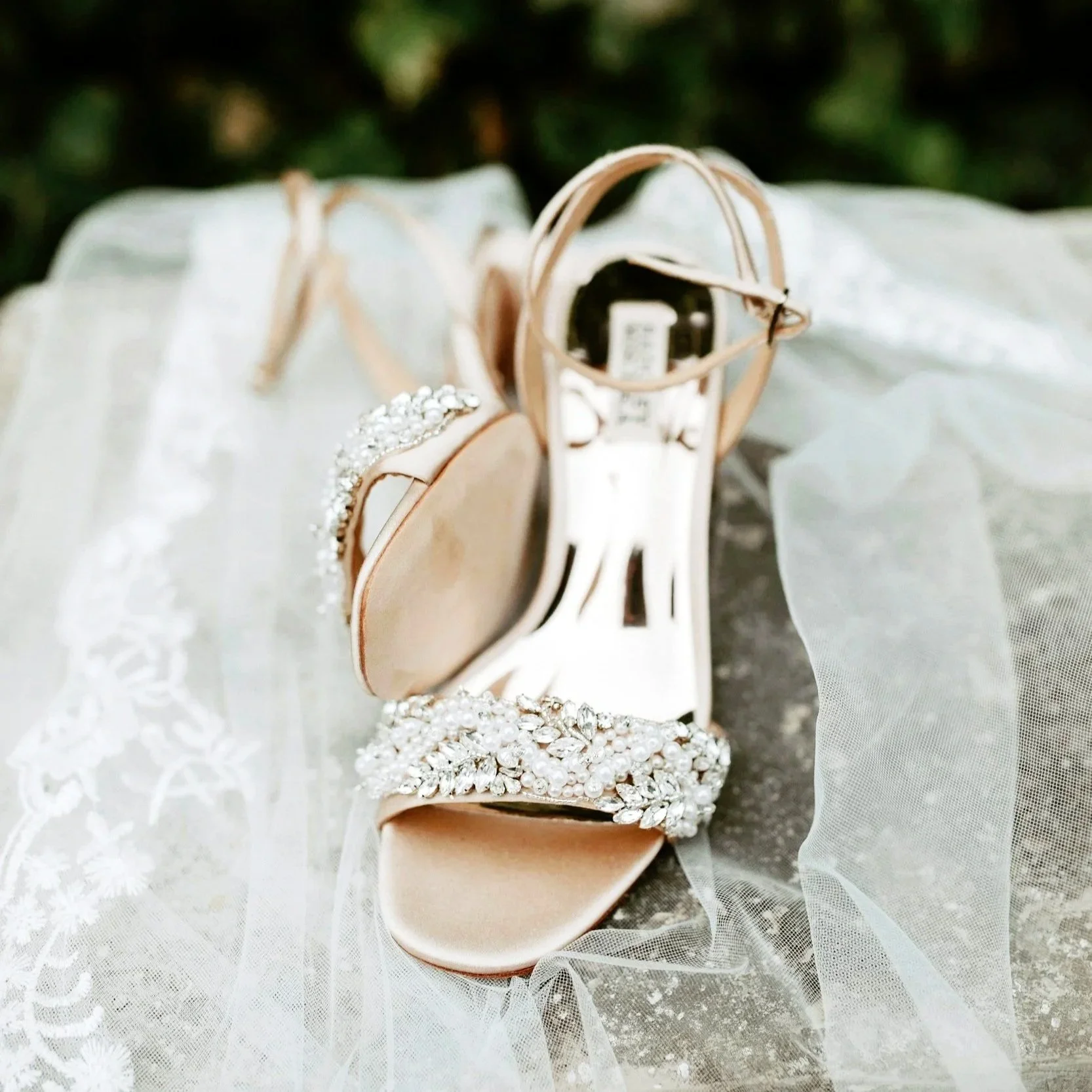 Elegant beige high-heeled shoes with rhinestone embellishments, placed on a lace fabric, alongside a small beige purse.
