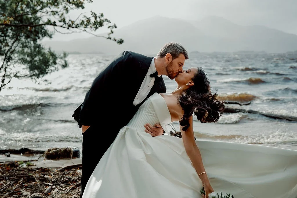 A newlywed couple sharing a kiss on a beach in Killarney National Park, Co.Kerry with the groom in a tuxedo and the bride in a white wedding dress.