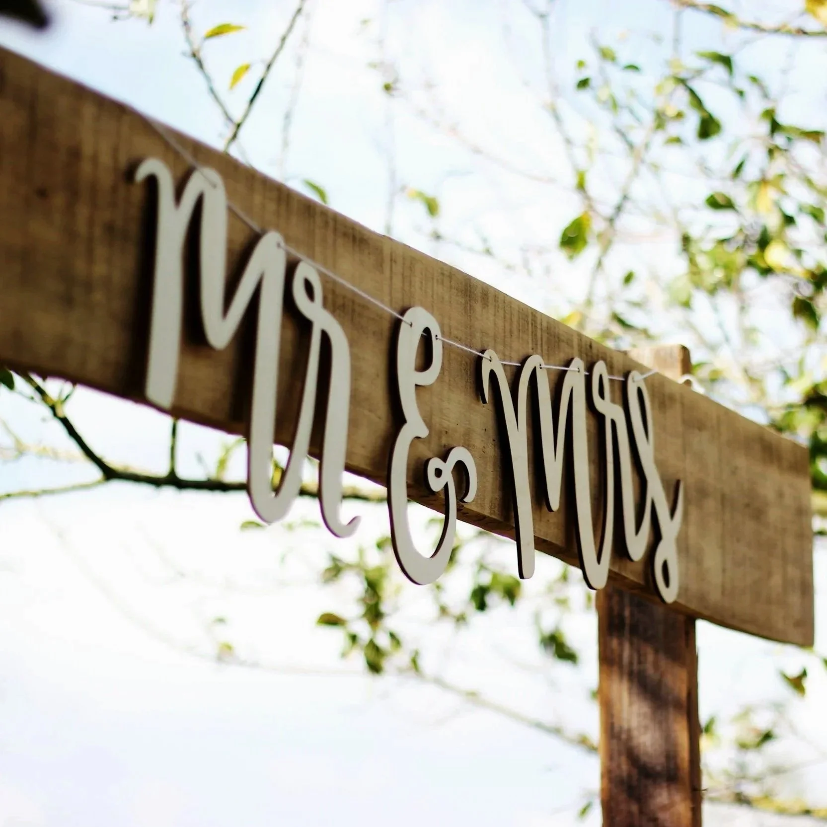 Wooden sign with white cut-out letters hanging on a line, reading "Mr & Mrs" on a tree with leaves and a blue sky in the background.