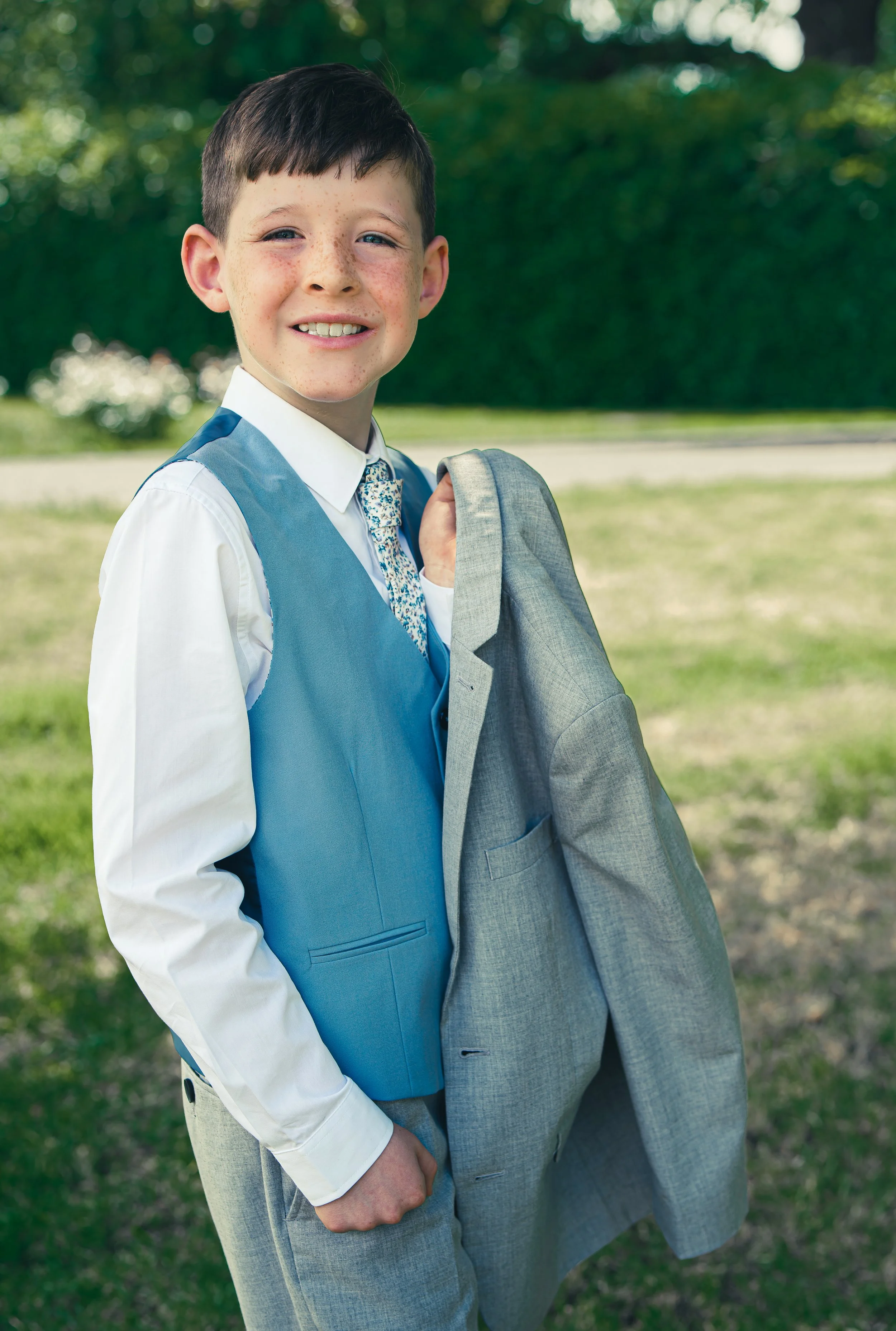 A young boy dressed in a white shirt, blue vest, and patterned tie, holding his gray suit jacket over his shoulder, smiling outdoors with green trees and grass in the background on his communion day.