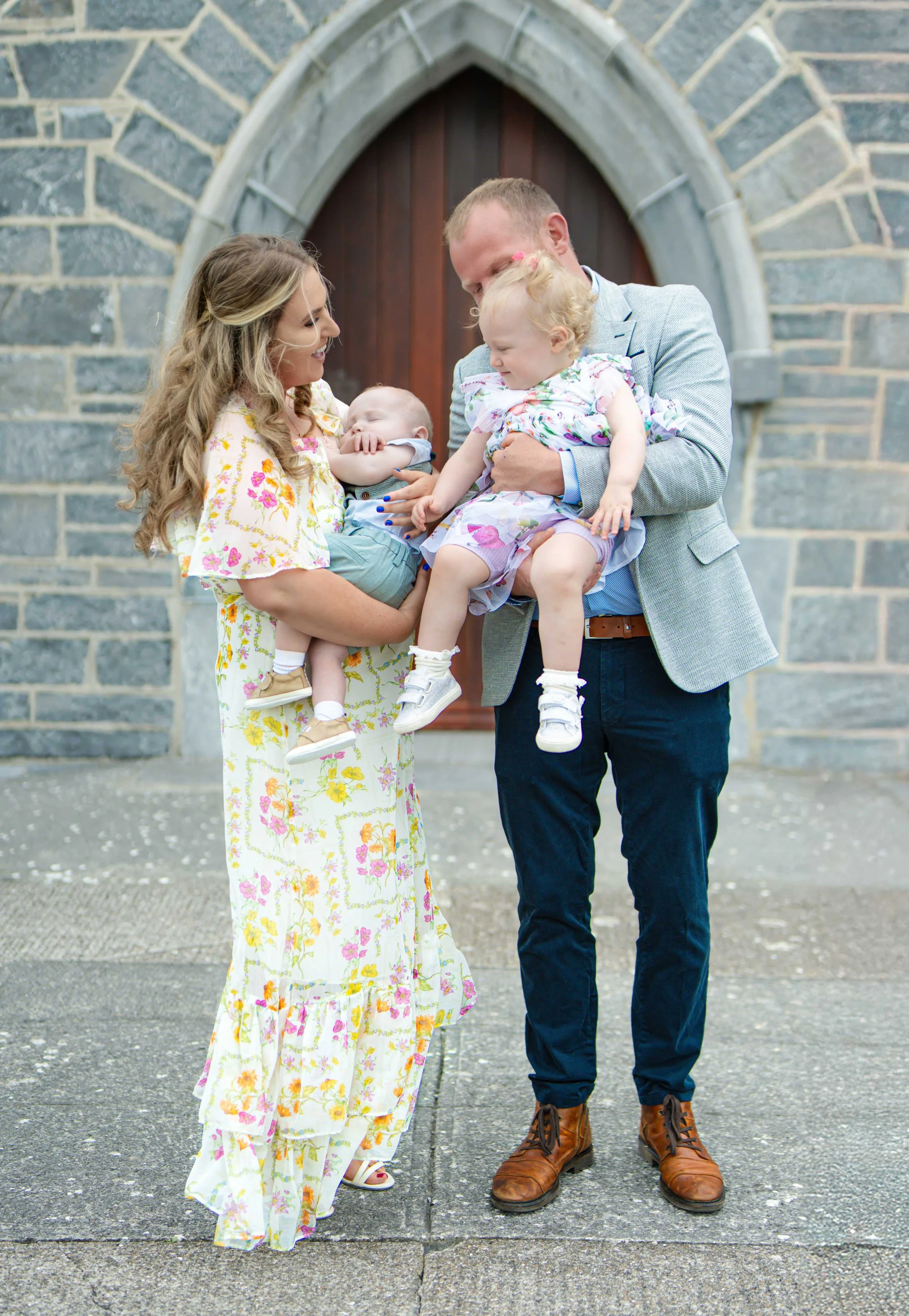 Family of four, a woman, a man, and two young children, standing outside Ardfert church. The woman is holding a baby, and the man is holding a toddler. They are smiling and looking at each other.