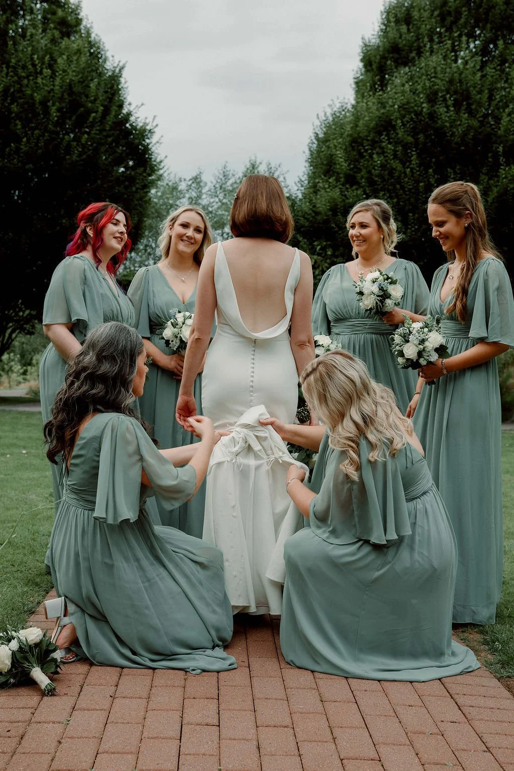 A bride in a white wedding dress surrounded by her bridesmaids in matching light blue dresses, outdoors on a cloudy day, with trees in the background.