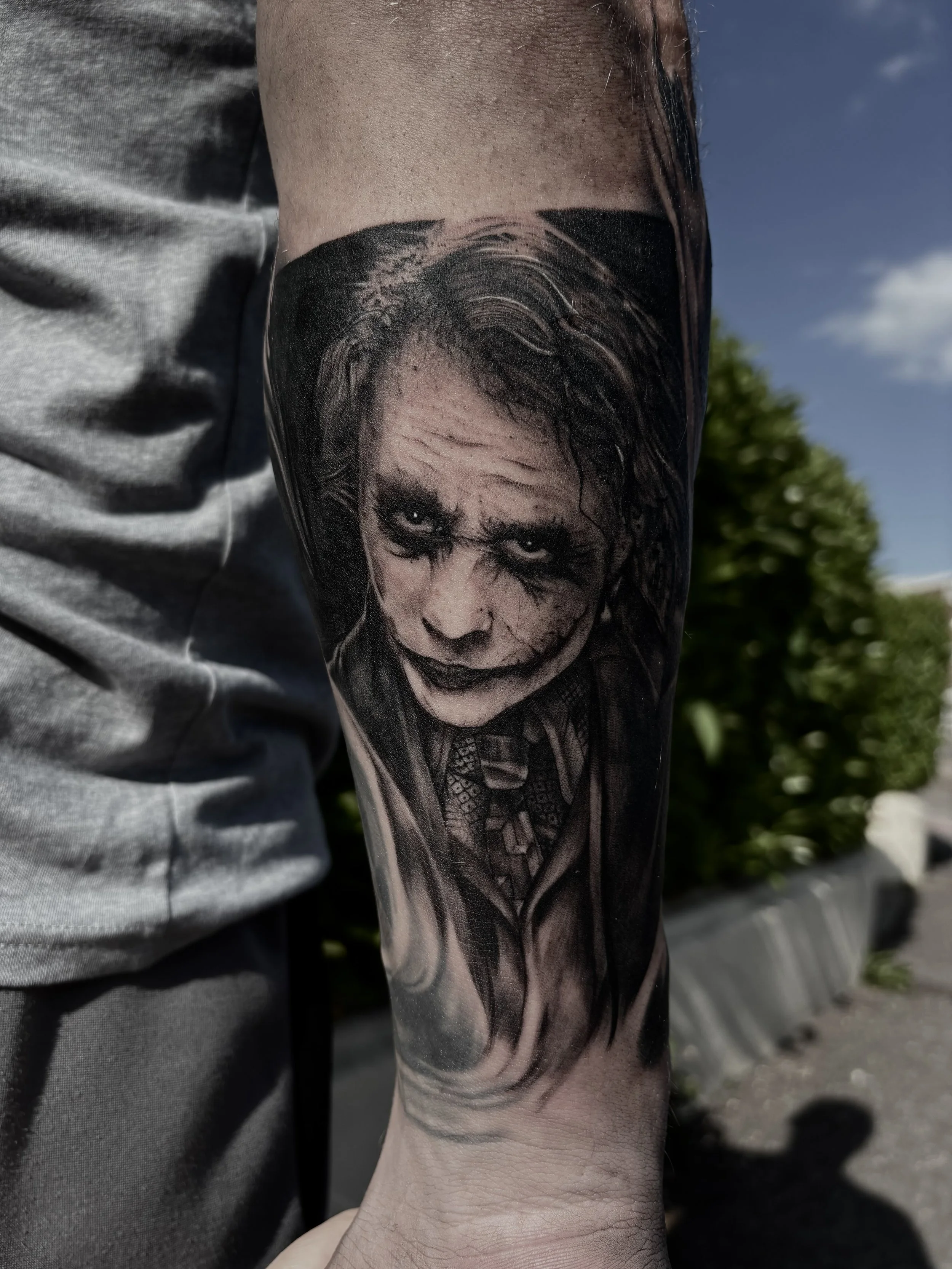 Black and gray tattoo of a face resembling the Joker from Batman, with white eyes, dark makeup, and a sinister expression, on a person's forearm.