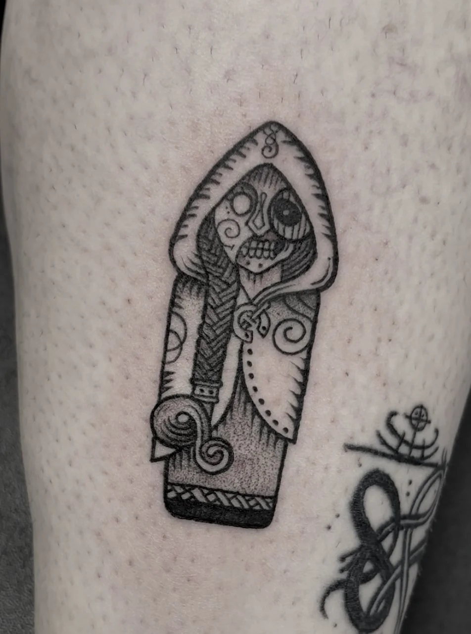 A black and gray tattoo of a stylized skeleton holding a sword, wearing a hooded cloak, with intricate line details, on light skin.