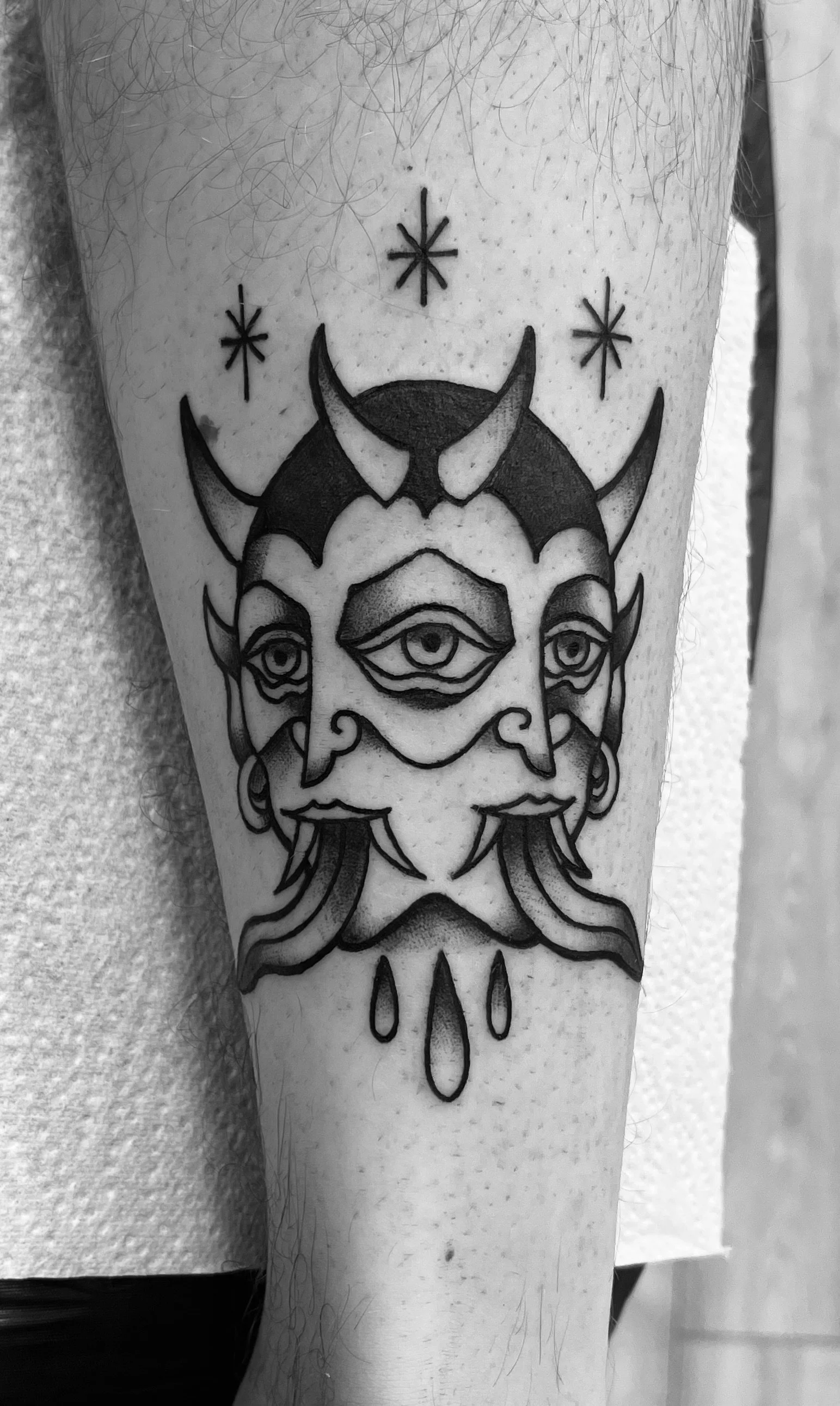 A tattoo of a mythological creature with three eyes, horns, and a beard, surrounded by celestial symbols and ink drops, on a person's forearm.