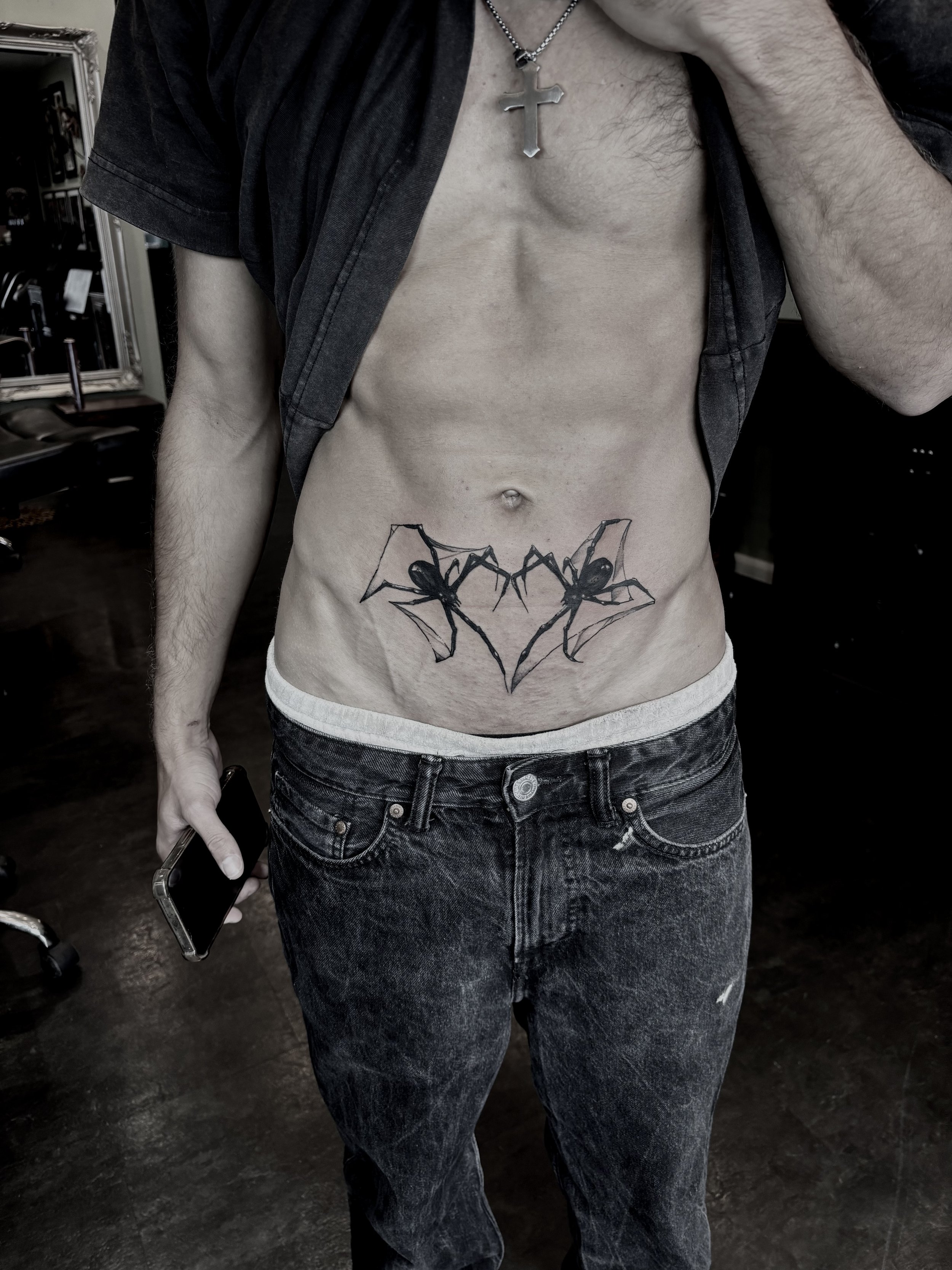 A man with a cross necklace taking a mirror selfie, showing a tattoo of two spiders on his lower abdomen.
