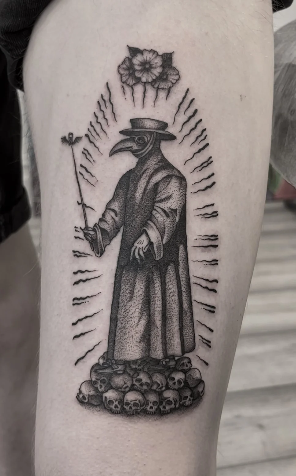 Tattoo of a plague doctor with a bird-like mask, wearing a long robe and wide-brimmed hat, holding a staff with a small winged figure, surrounded by radiating lines, standing on a pile of skulls, with flowers above.