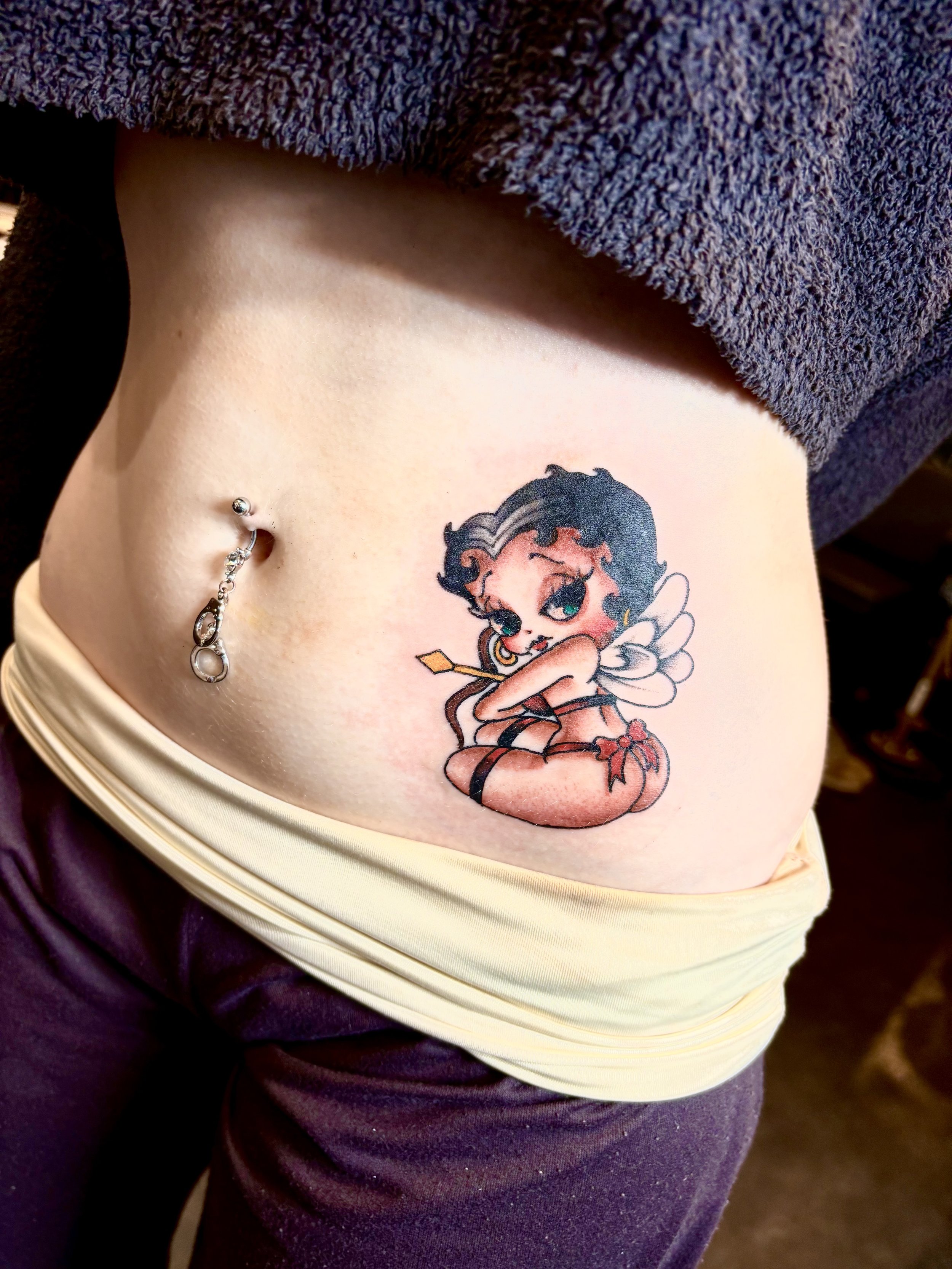 Tattoo of Betty Boop with fairy wings and a bow on her head on a person's side torso.