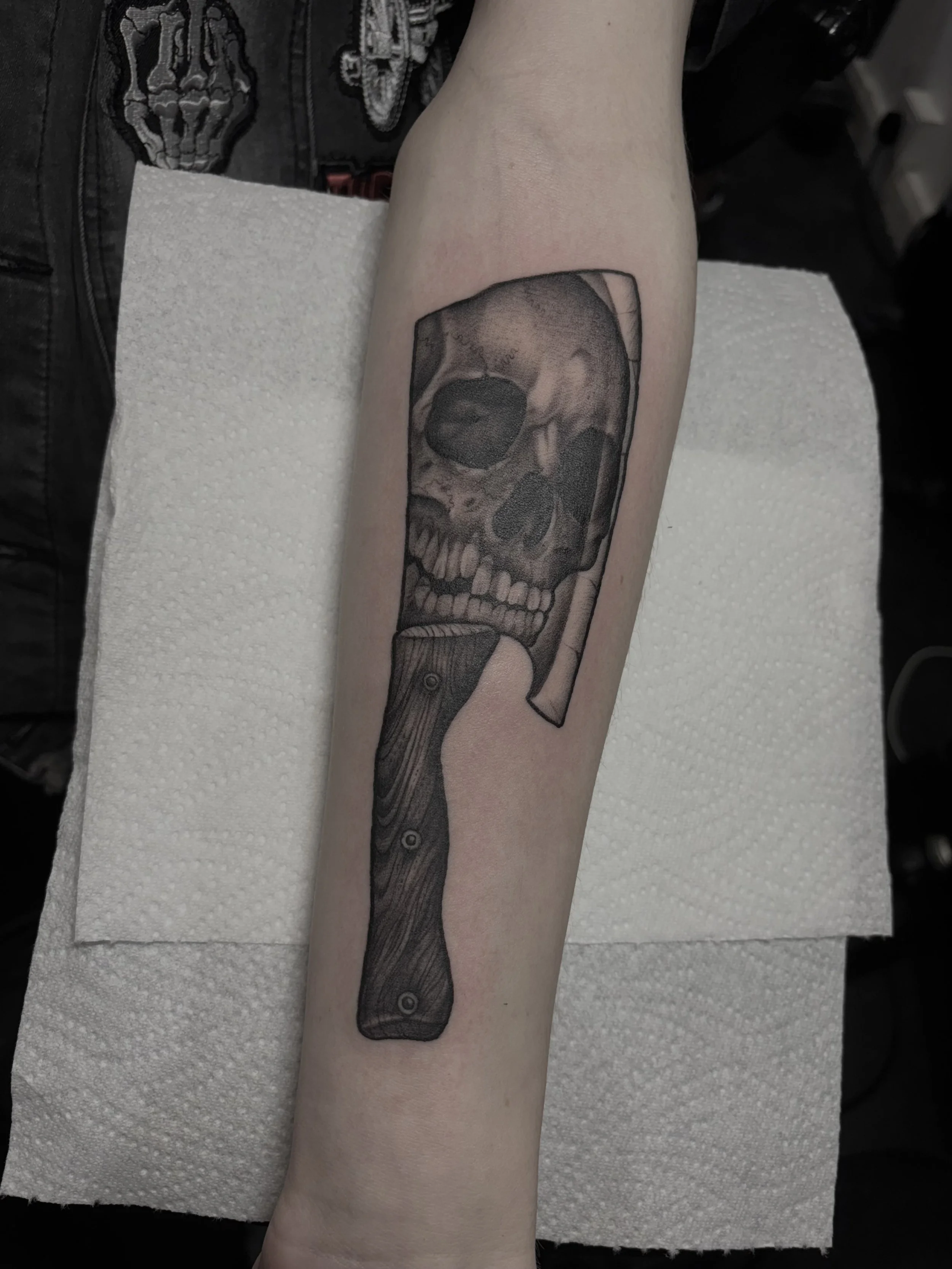 A tattoo of a human skull with a knife through the top of it, located on a person's forearm.