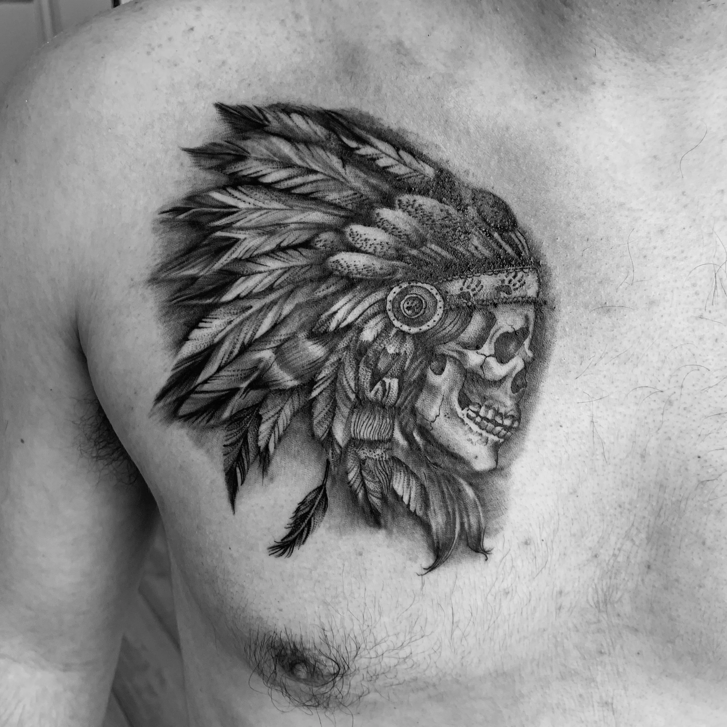 Black and white tattoo of a human skull wearing a Native American headdress of feathers on a person's chest and shoulder