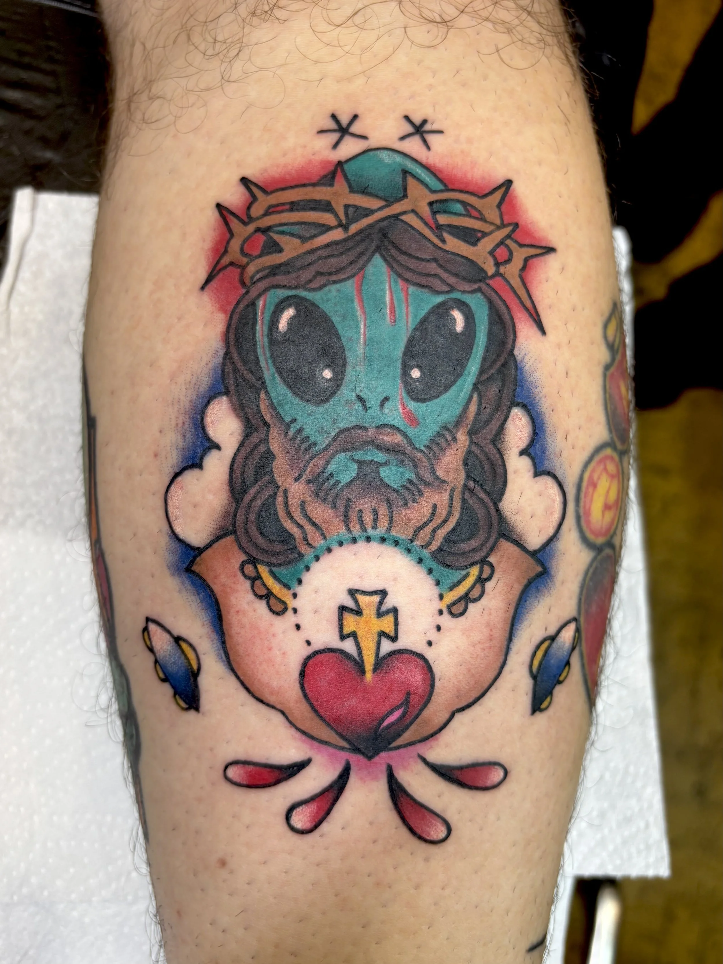 Tattoo of a cartoon-style alien with big black eyes, wearing a crown of thorns, and with a beard, depicted as Jesus Christ with a heart and cross at the bottom.