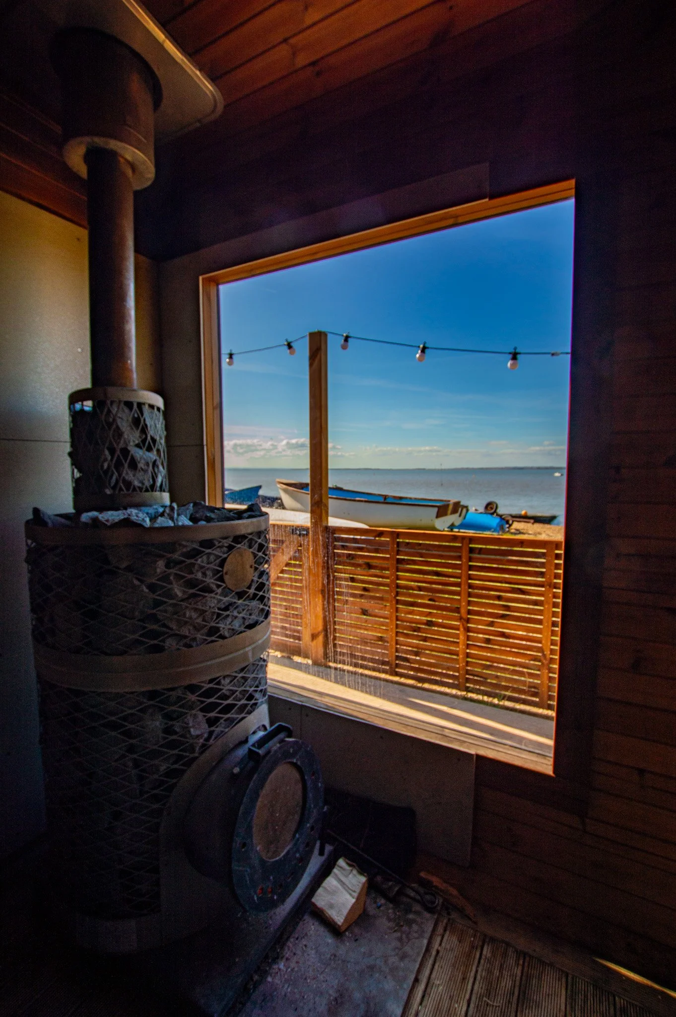 View from inside a wooden room looking out a large open window onto a beach with boats, a fence, and string lights overhead, with a wood stove in the foreground.