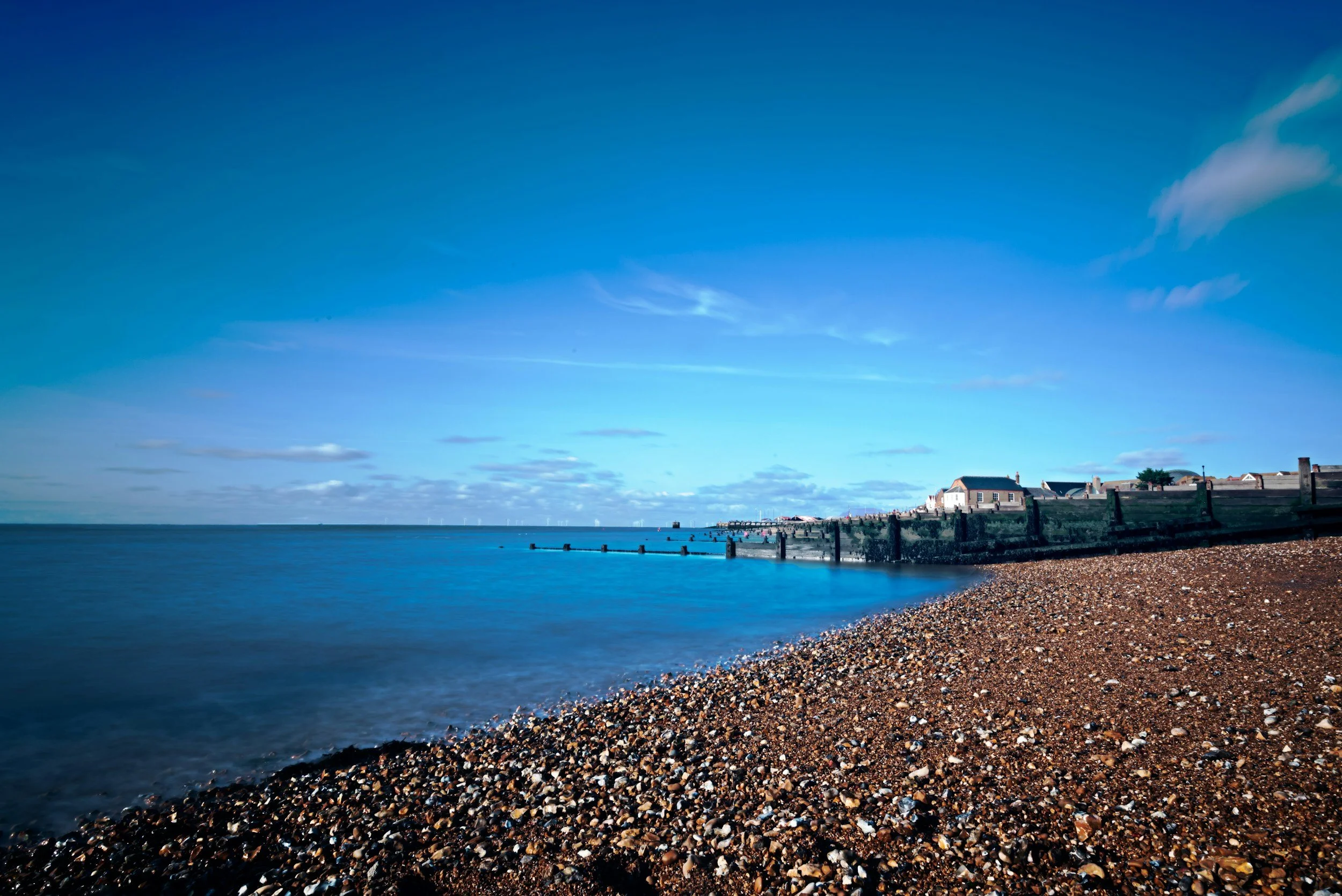 A rocky beach with a view of a calm blue sea and a small harbor with buildings in the distance under a mostly clear sky.
