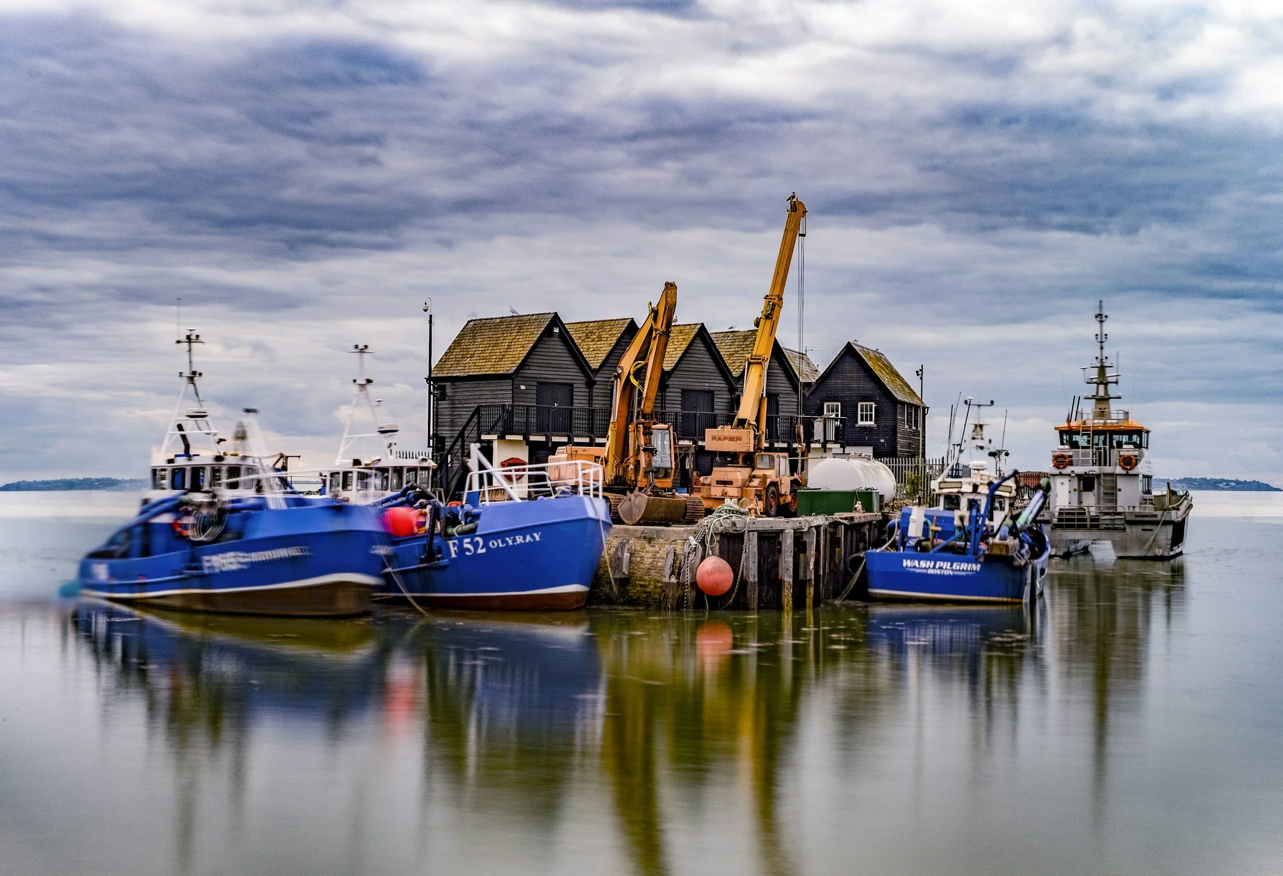 A small harbor with three boats docked next to wooden buildings, construction equipment on the pier, and a cloudy sky overhead.