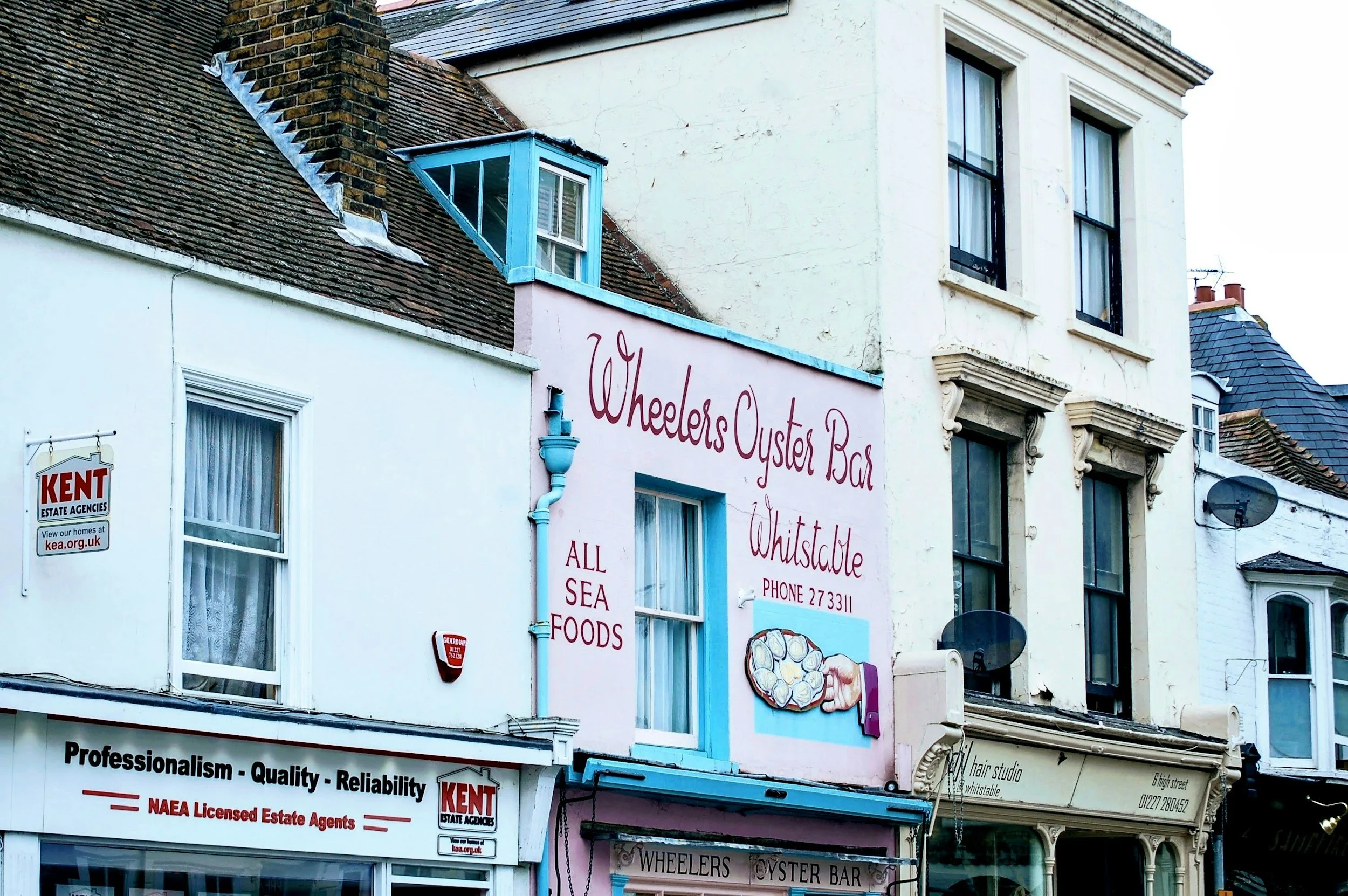 Facades of multiple buildings on a street, including a sign for Wheeler's Oyster Bar, which features a painting of oysters and a phone number. The buildings have various signs, windows, and architectural details, with a mix of pastel and neutral colors.