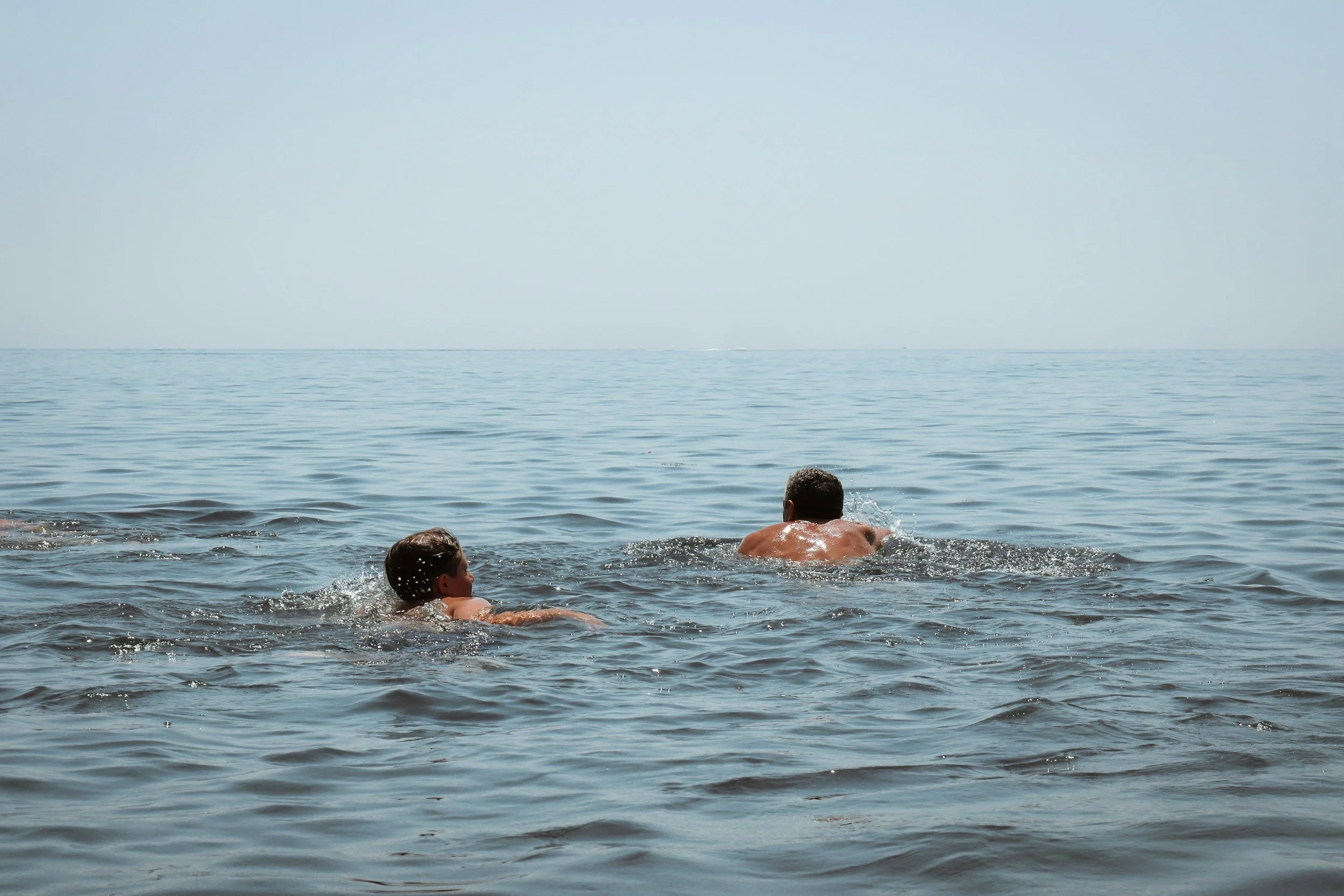 Two people swimming in the ocean, one adult male and one young boy, with calm water and a clear sky.
