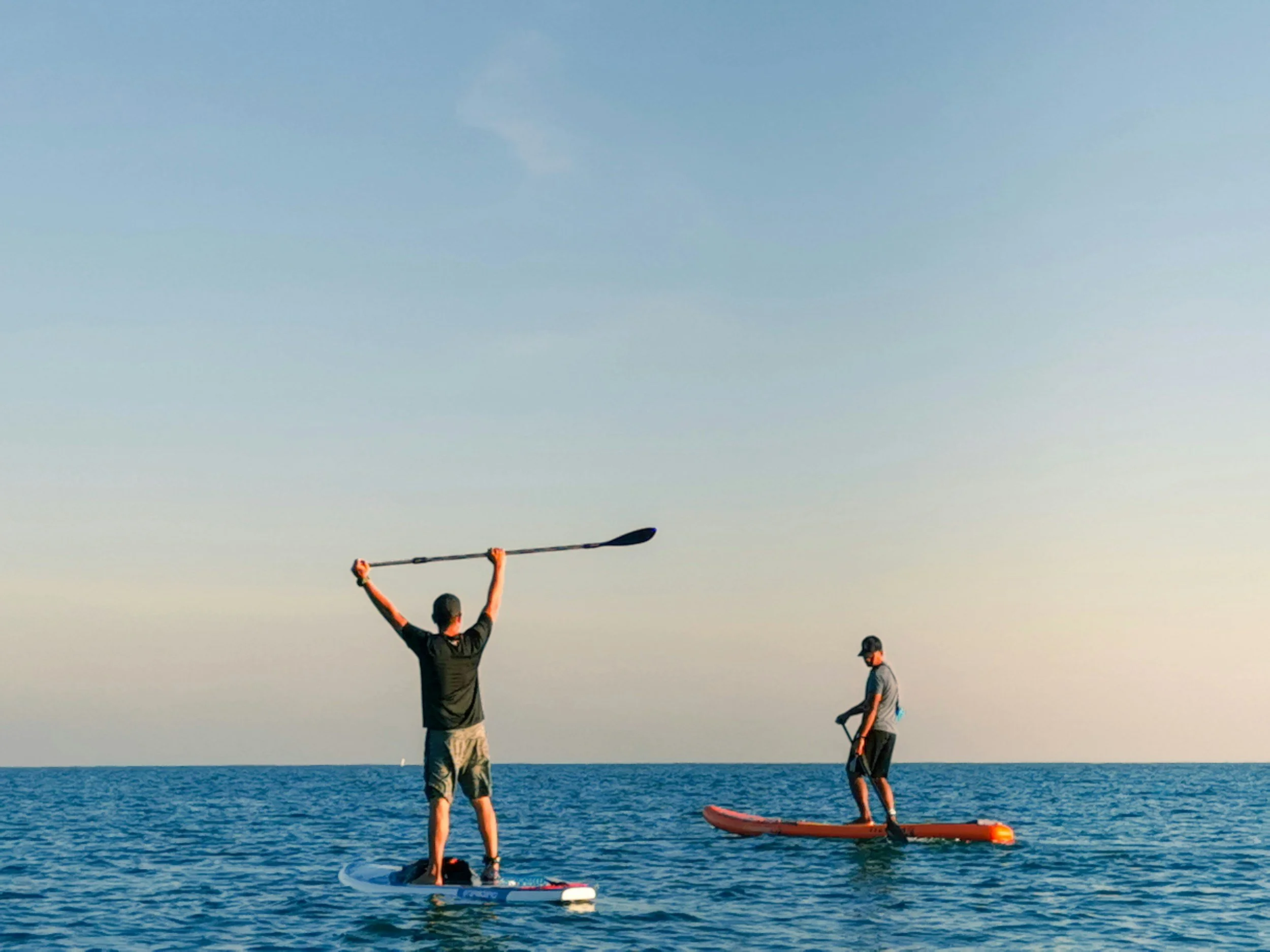 Two people paddleboarding on the ocean with clear skies in the background.