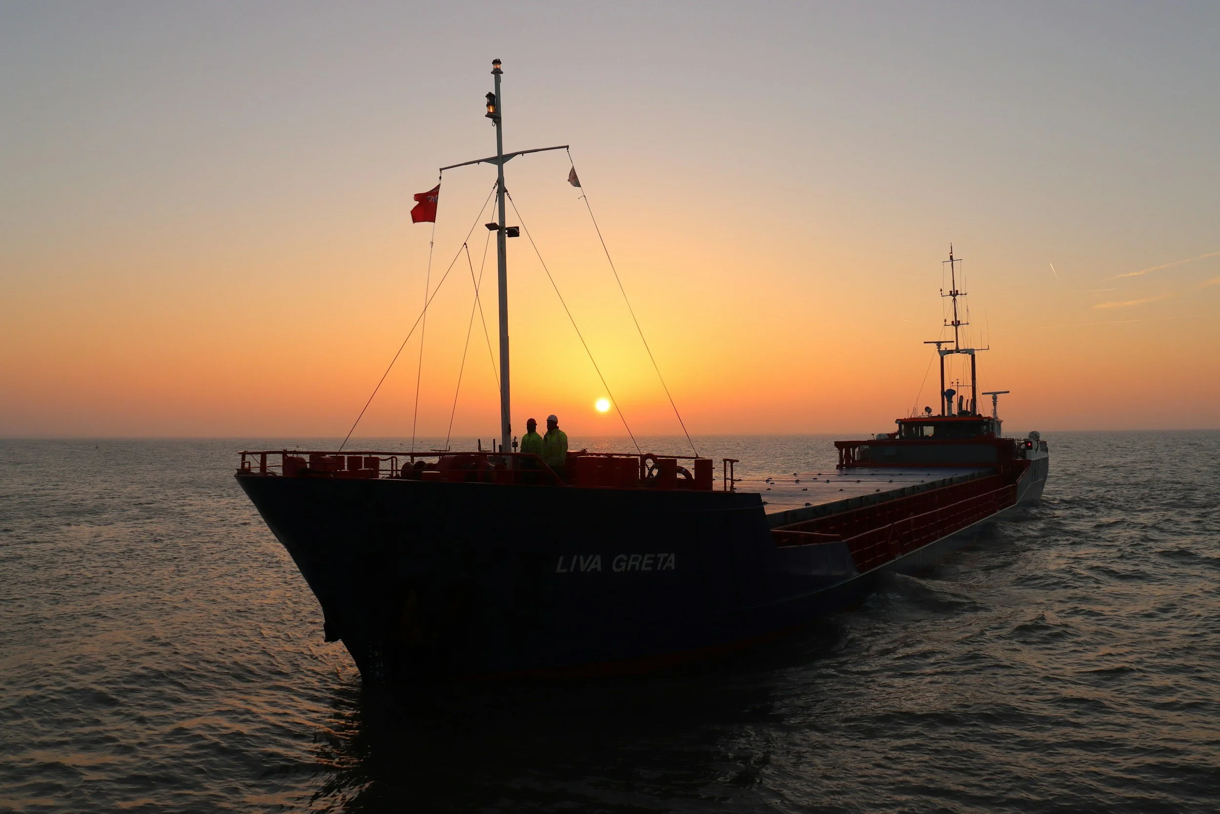 A cargo ship named LIVA GRETA on the water at sunset with two people on deck and the sun setting in the background.