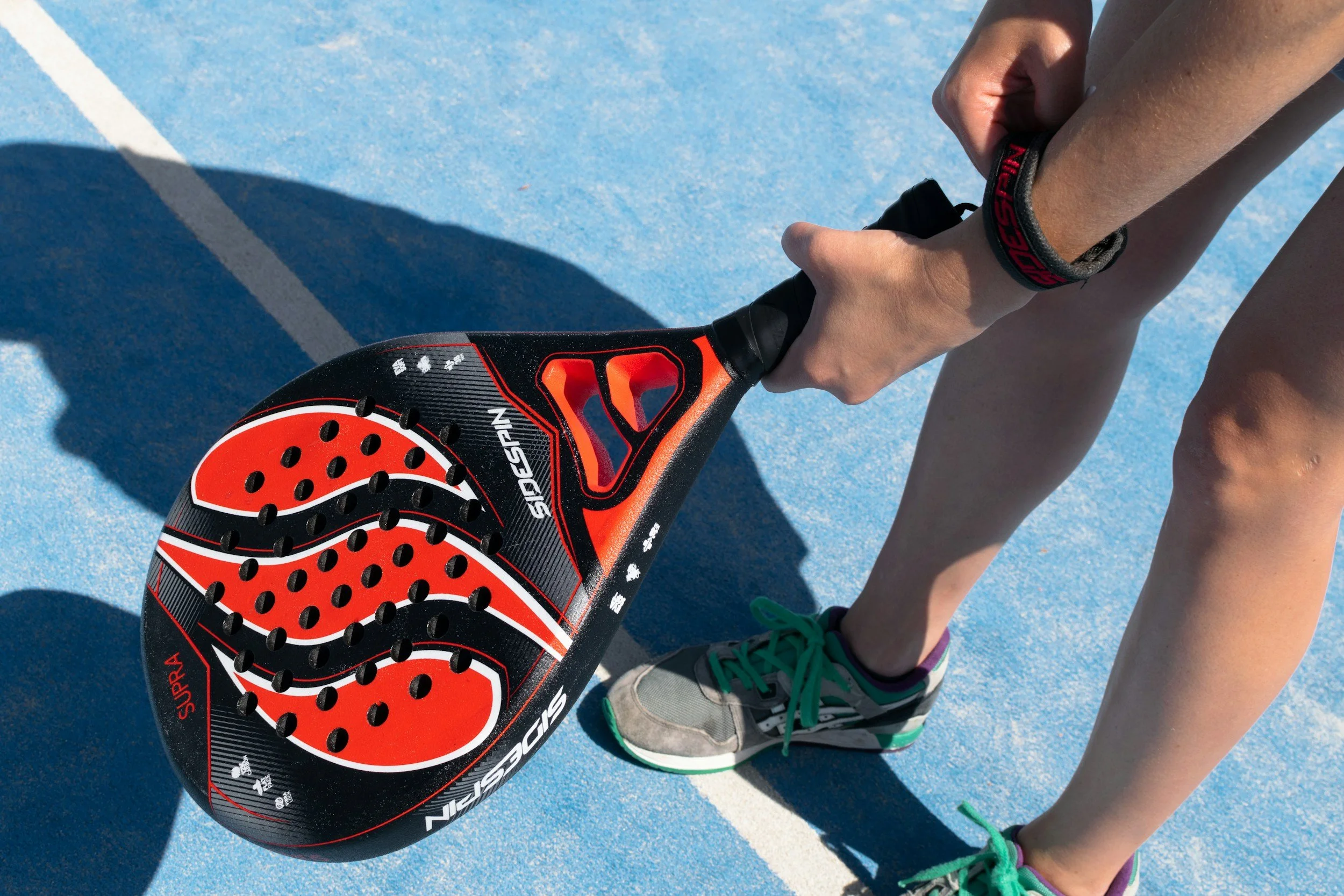 A person holding a black paddle with red and white accents on a blue outdoor sports court, wearing athletic shoes and a watch.