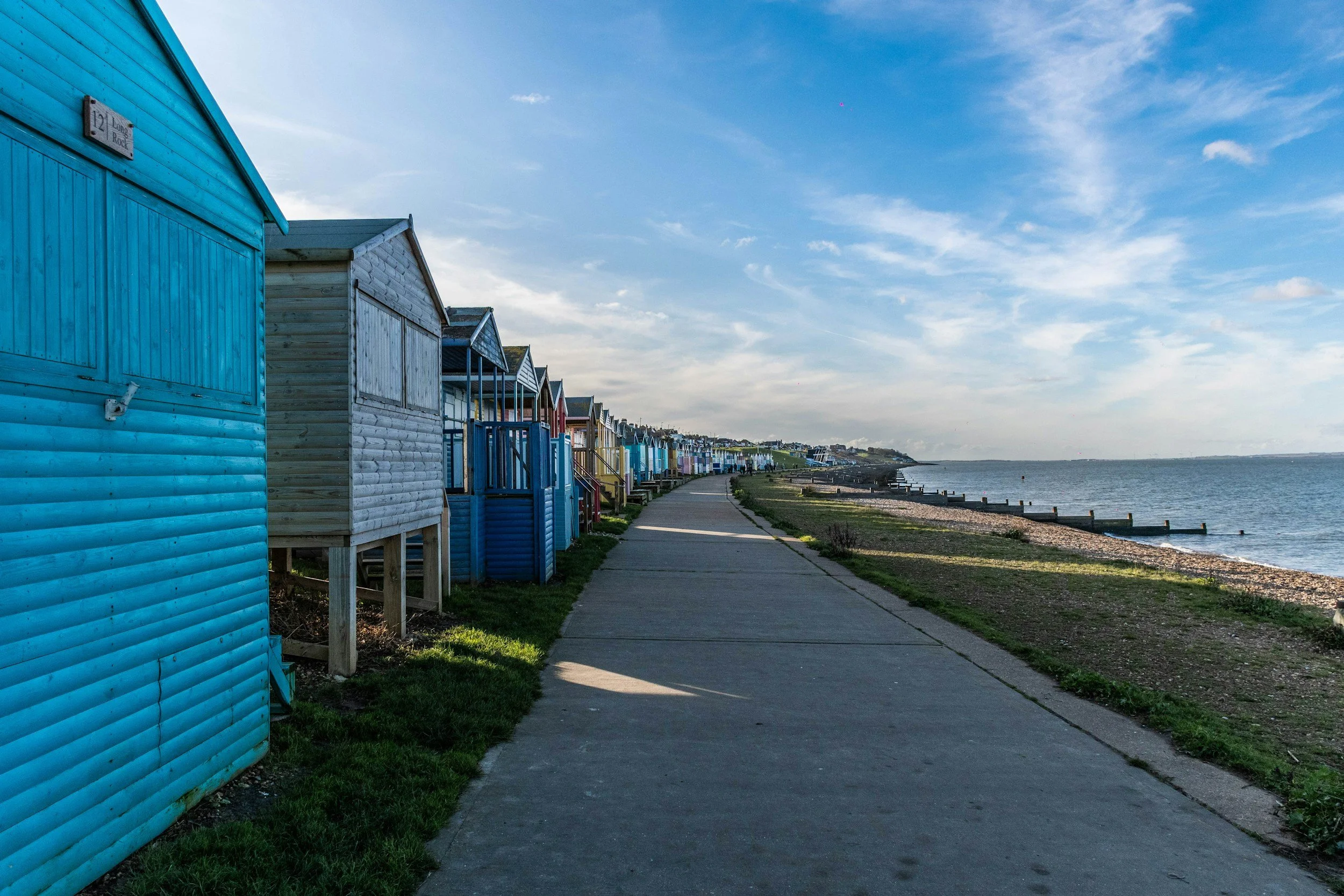 Colorful beach huts lining a paved pathway along a coastline under a partly cloudy sky.