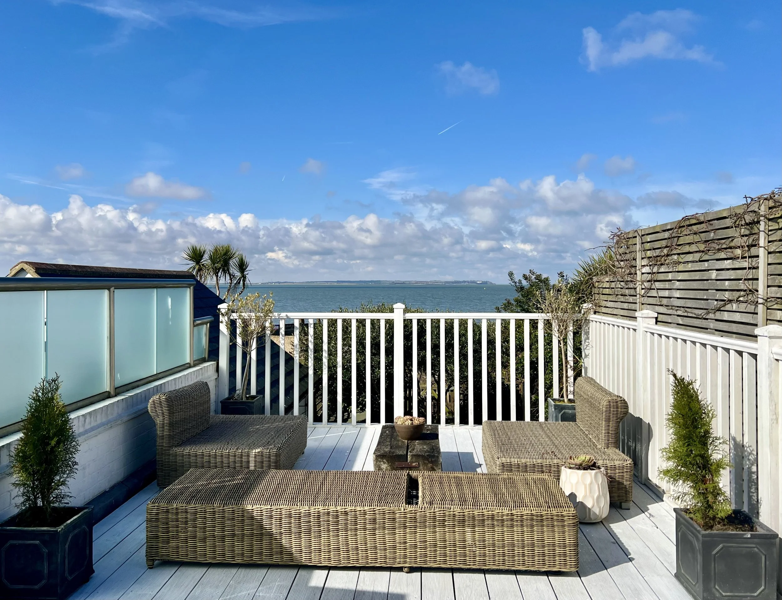 Outdoor balcony with wicker furniture, potted plants, and a view of the ocean and blue sky with clouds.