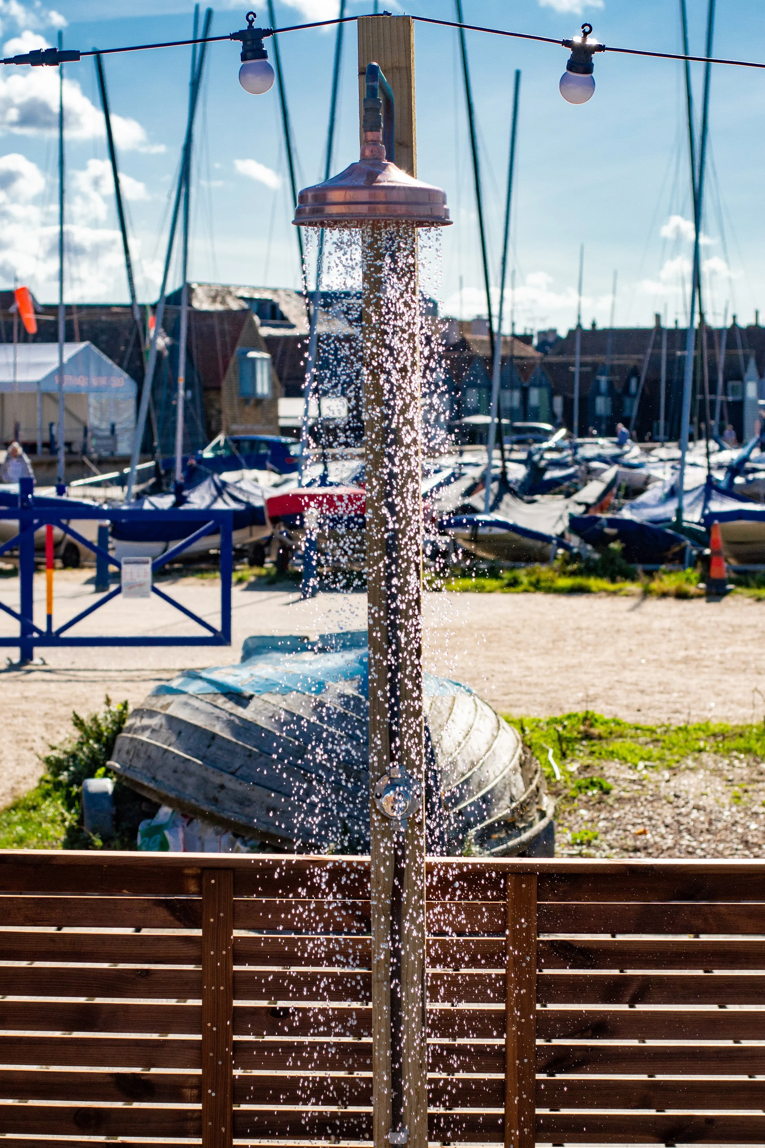An outdoor shower with water flowing, located near a marina with boats and sailboats in the background, under a partly cloudy sky.