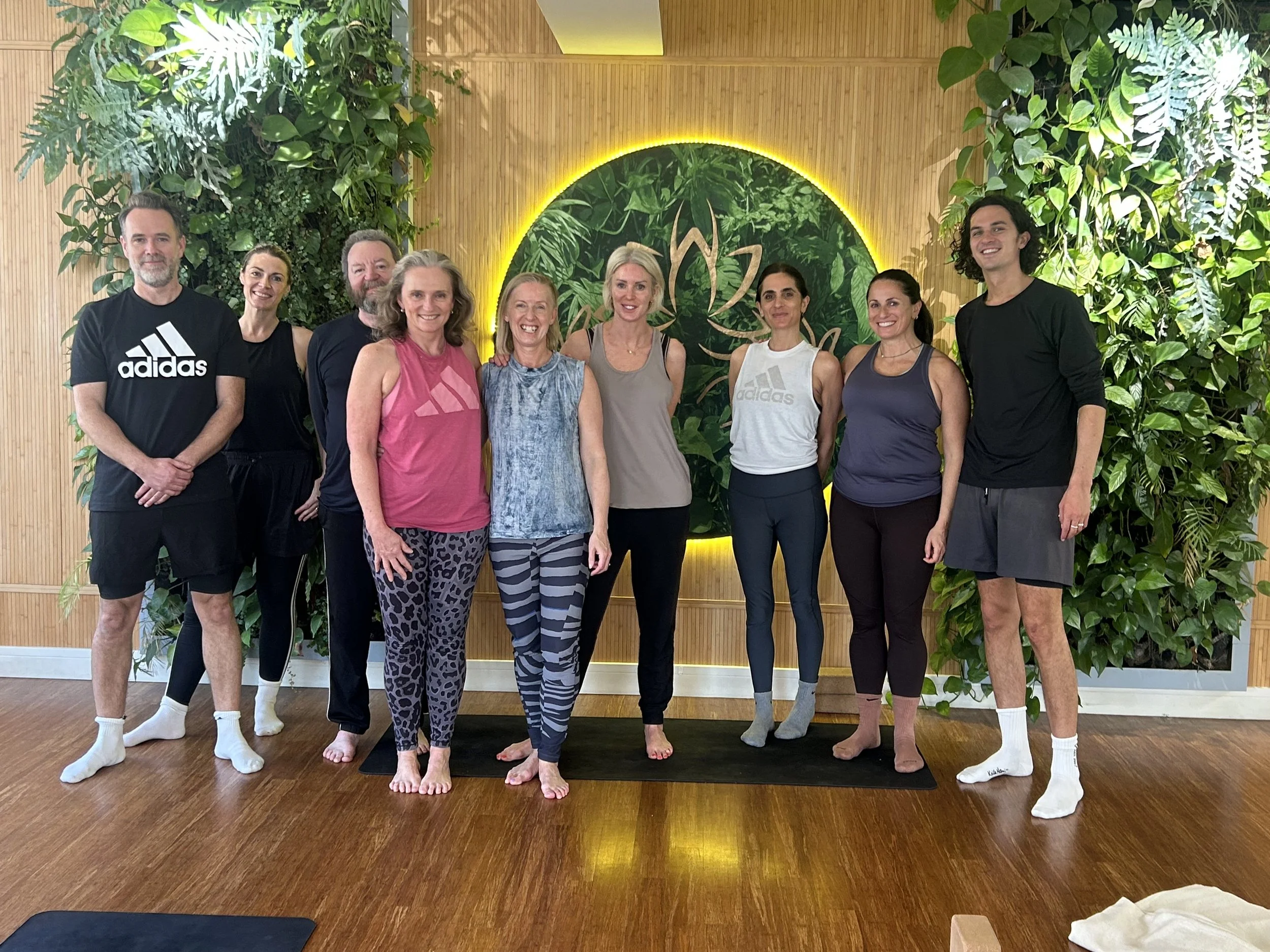A group of nine people standing on yoga mats in a room with wooden floors and green plant decor, smiling at the camera during a yoga class or group session.