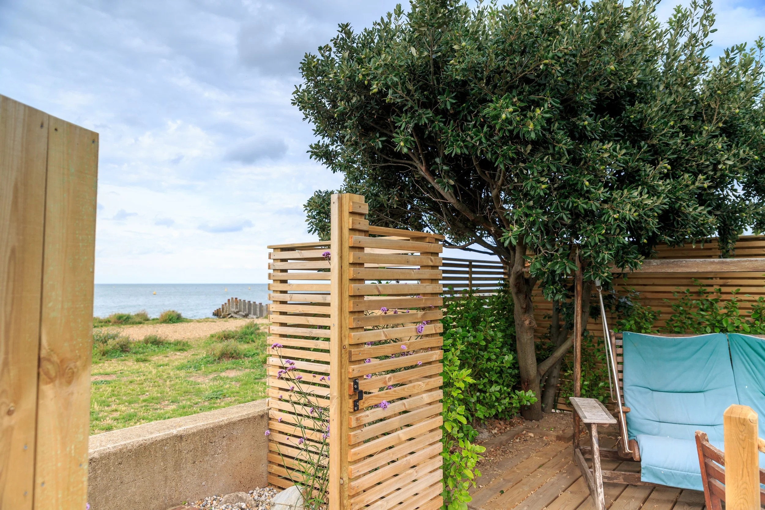 A cozy outdoor patio area with a wooden fence, a large leafy tree, a blue cushioned outdoor chair, and a view of the beach and ocean in the background.
