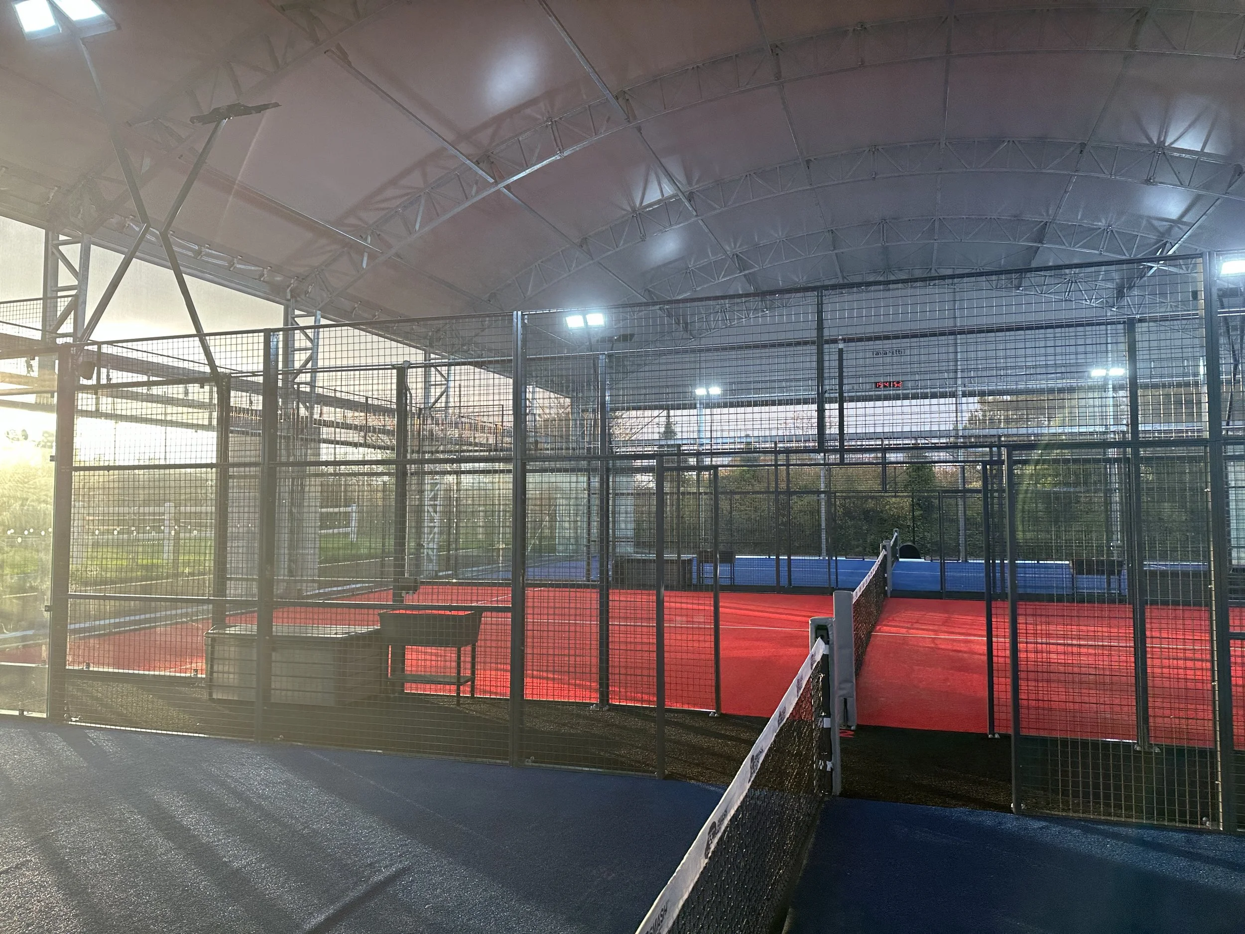 Indoor padel court with red playing surface, surrounded by black metal fencing, with overhead lighting and a blue court in the background.