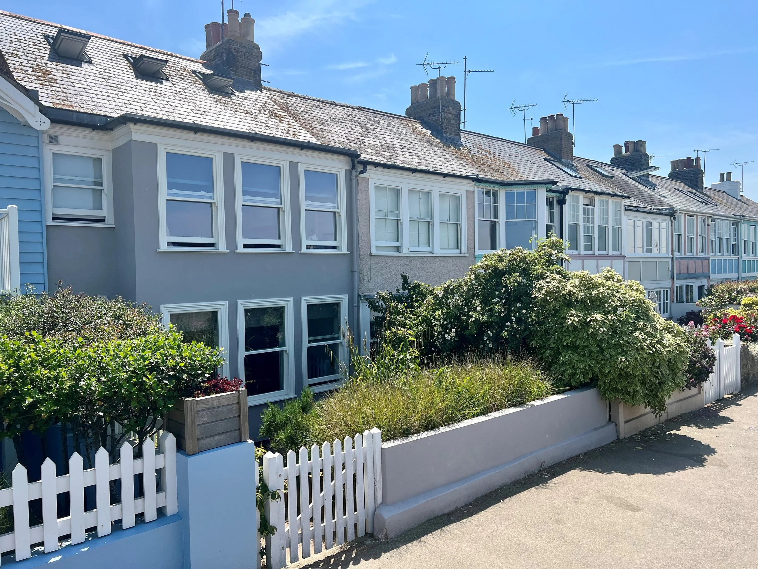 Row of colorful seaside houses with white picket fences, lush bushes, and clear blue sky.