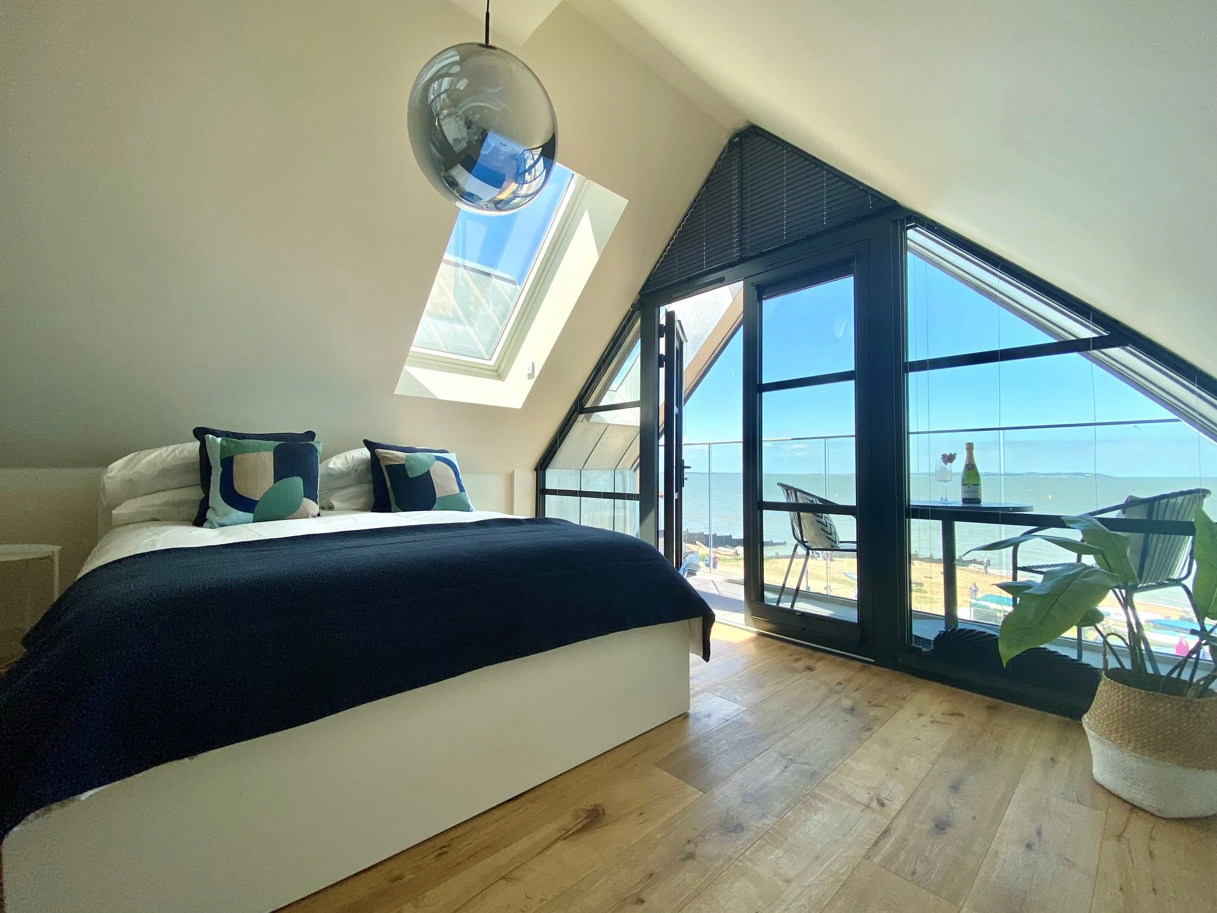 A bedroom with a bed, pillows, and a black bedspread. There are sliding glass doors leading to a balcony with ocean views. The room has a sloped ceiling with two skylights, and a hanging spherical light fixture.
