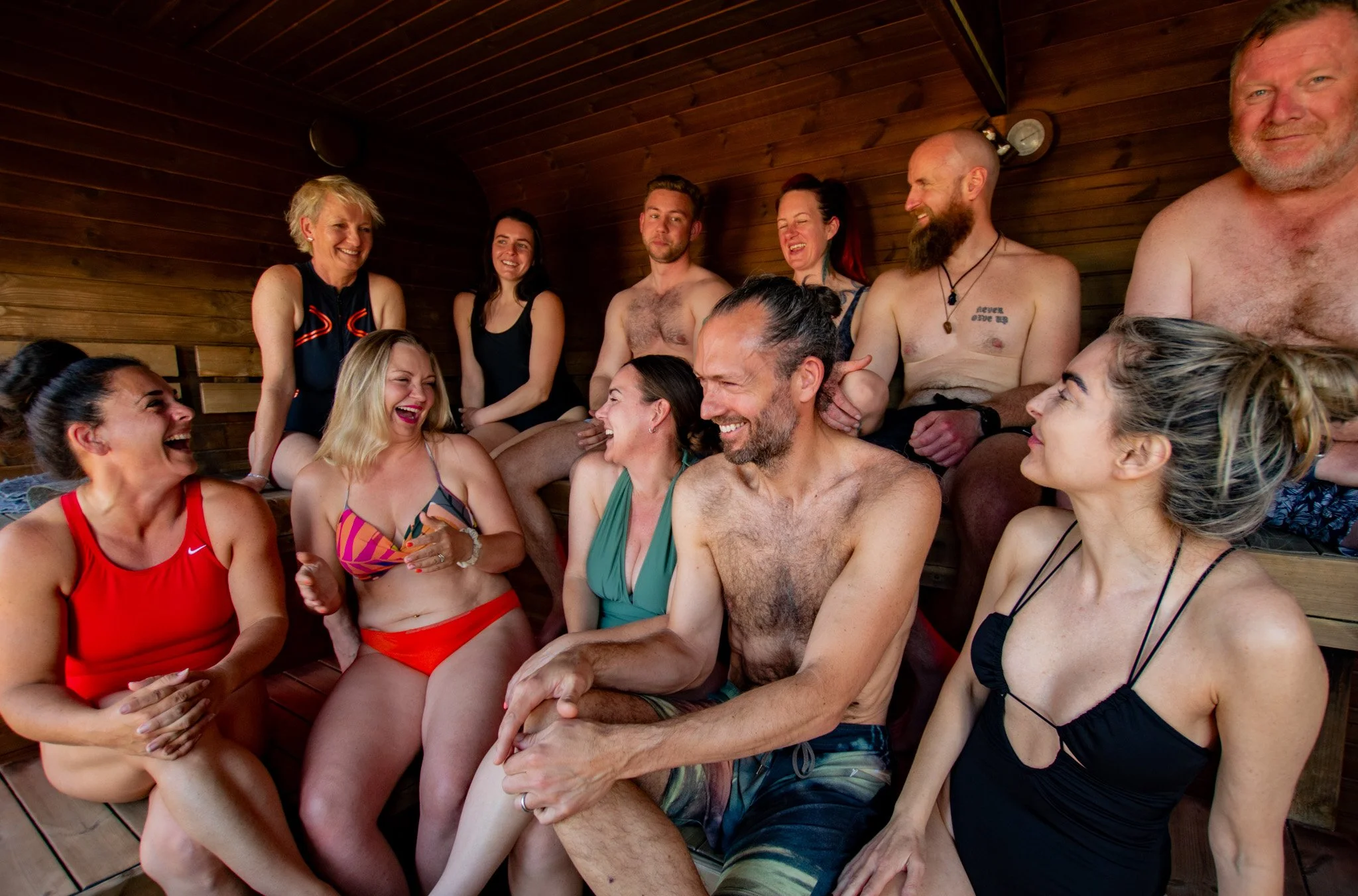 A group of people laughing and talking inside a wooden sauna, wearing swimsuits, some are shirtless, and they appear to be enjoying each other's company.