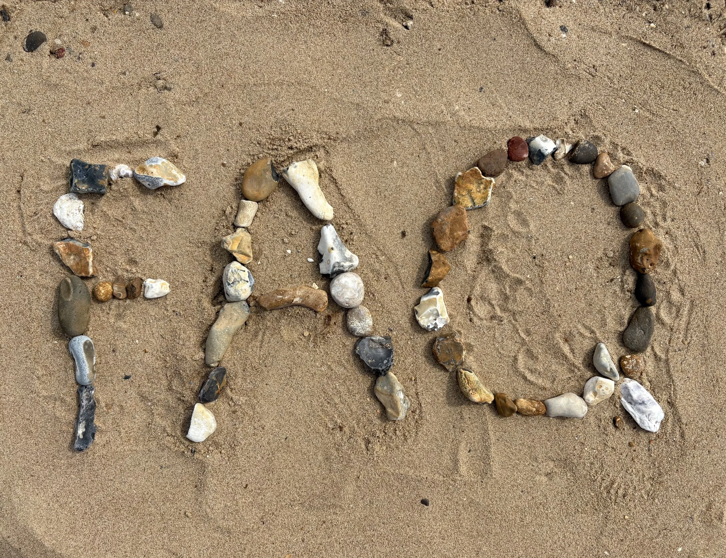 The image shows the word 'FAQ' written on sandy ground using various small rocks and pebbles.