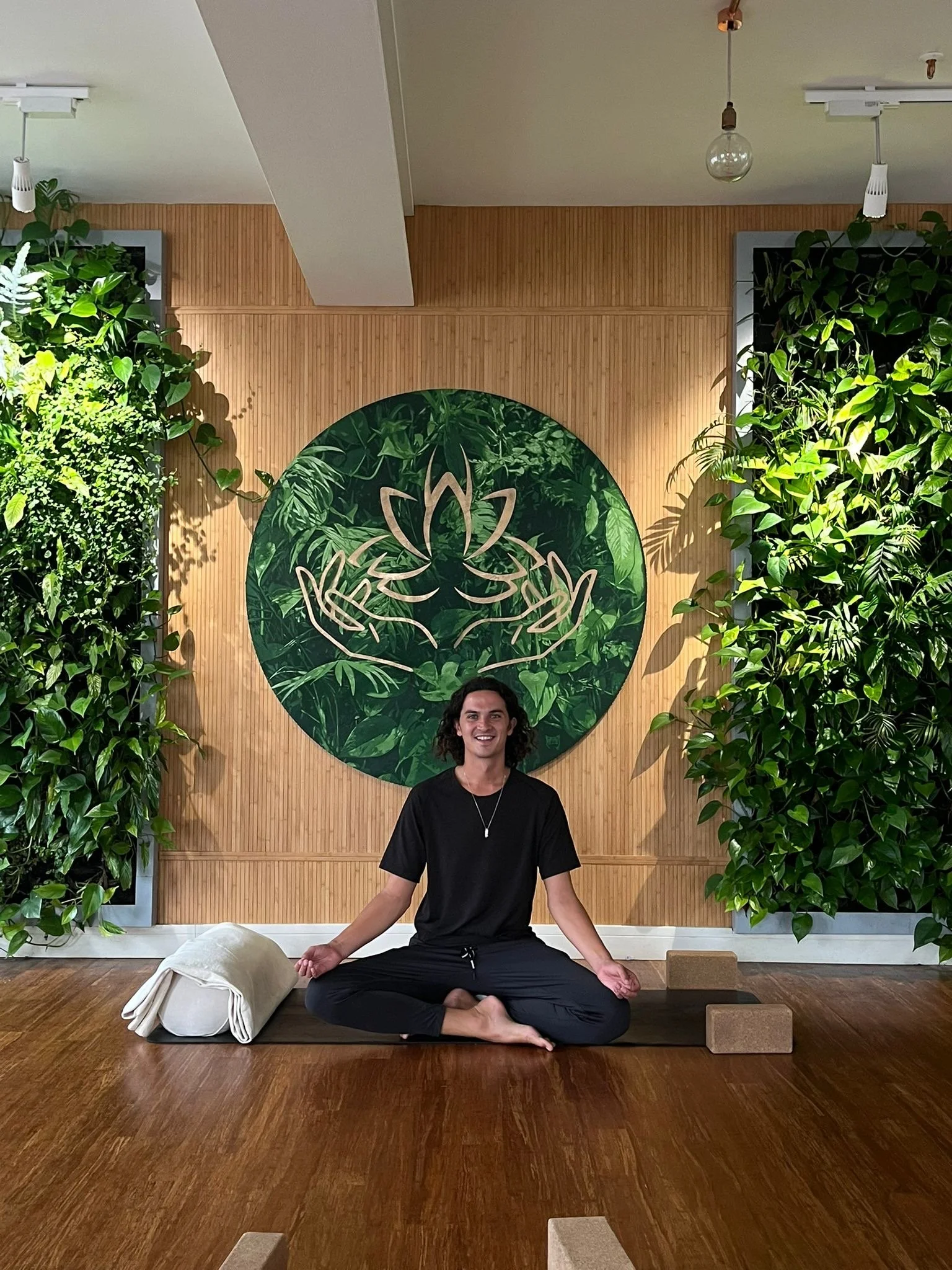 A person sitting cross-legged on a yoga mat in a meditation pose in front of a wooden wall with a large circular green mural featuring a stylized lotus flower and leaves, surrounded by green plants.