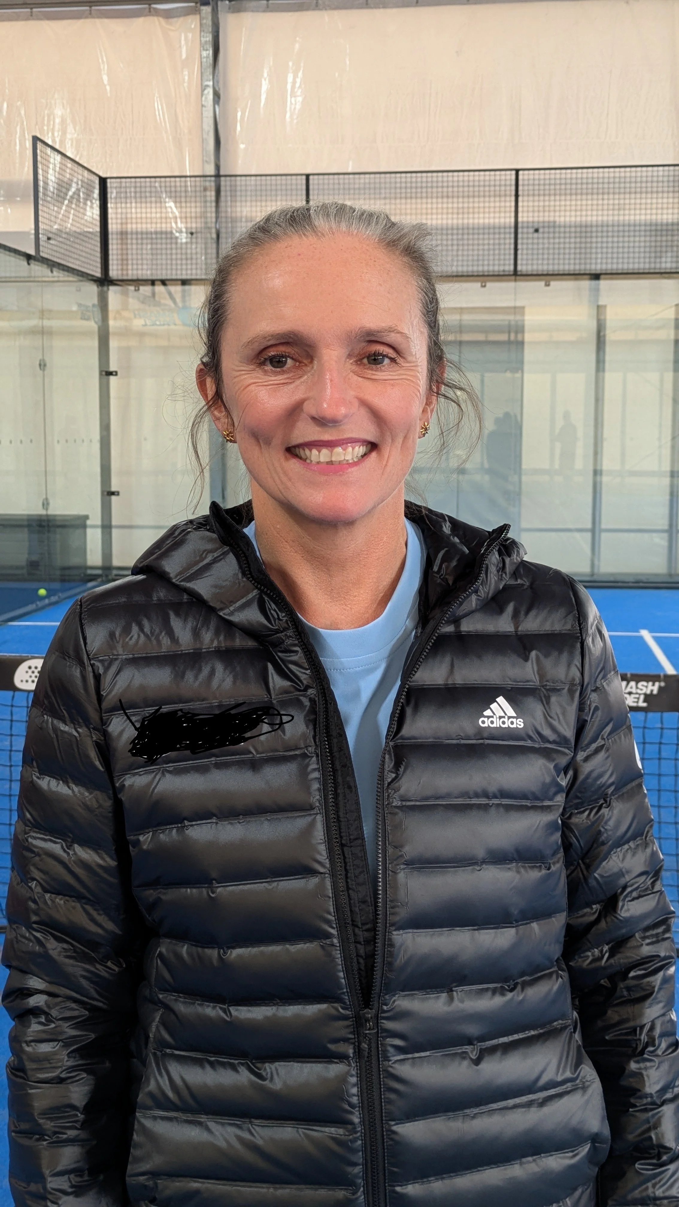 A woman smiling, standing in front of a tennis court.