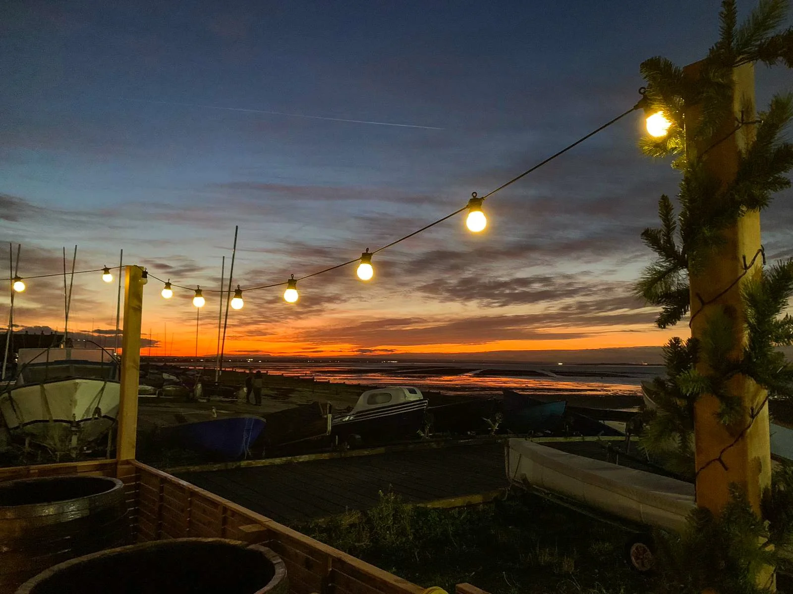 Sunset over a coastal area with boats on the shore, illuminated string lights hanging, and a tree with string lights wrapped around its trunk.