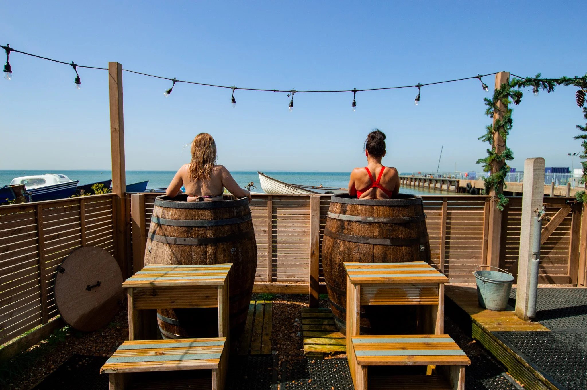 Two women sit in wooden barrel hot tubs outdoors by the seaside, facing the ocean on a sunny day, with a fence, small tables, and string lights overhead.