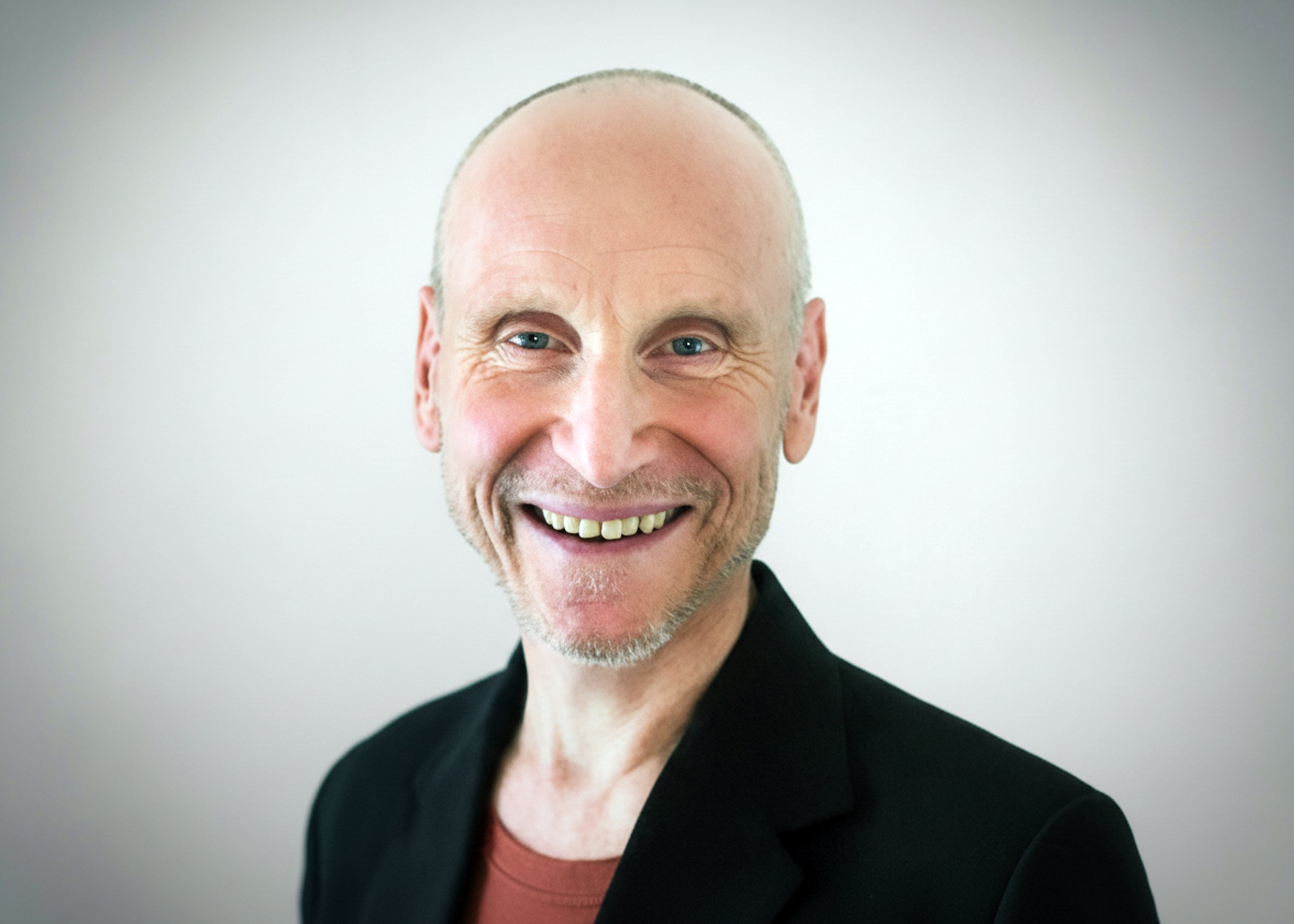 Portrait of a smiling, bald man with blue eyes, a light beard, wearing a black blazer and a reddish-brown shirt against a plain light background.