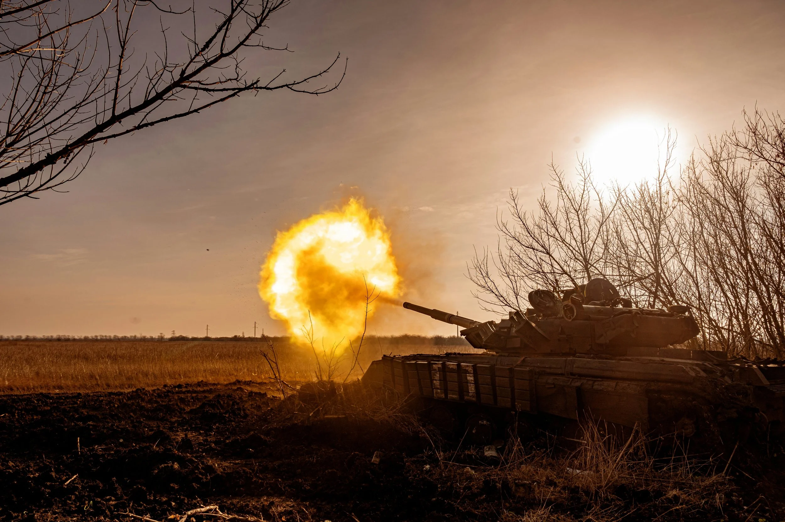 Tank firing a burst of flame and smoke in an open field with a bright sun and leafless trees in the background.