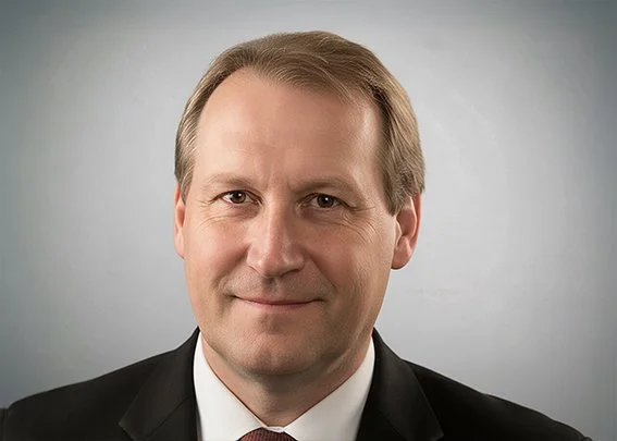 A professional headshot of a middle-aged man with light brown hair wearing a black suit, white shirt, and red tie, against a gray background.